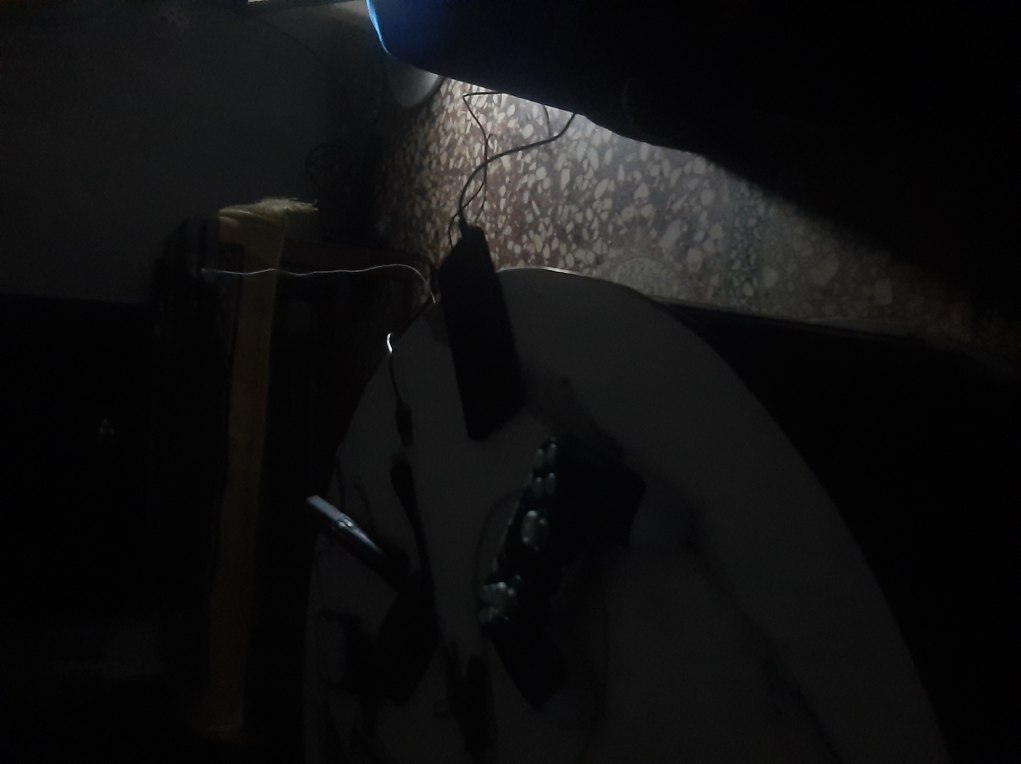 In a dimly lit room, a white round table is positioned in the foreground. On the table, a black gaming controller with multiple buttons is visible. To the left of the controller, a black object, possibly a phone or remote, lies on the table. A white cable snakes from the table towards the background. In the background, a dark surface with a textured pattern, likely a floor, is visible. A yellow object is partially seen behind the table. A person's dark clothing is present on the right side of the image.