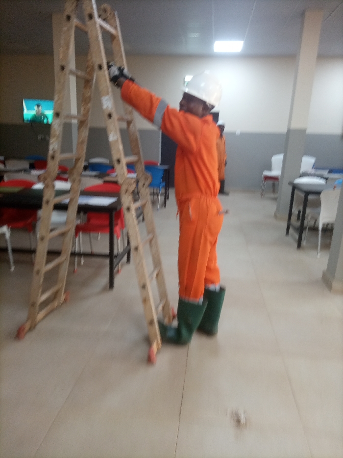 A person wearing an orange jumpsuit, white hardhat, gloves, and green rubber boots stands next to an A-frame ladder. The ladder is wooden and stands in a tiled room. The person is reaching up, appearing to adjust or inspect the ladder. In the background, there are tables and chairs, a television screen, and a support column. The lighting in the room is bright, possibly fluorescent. The image is slightly blurry.