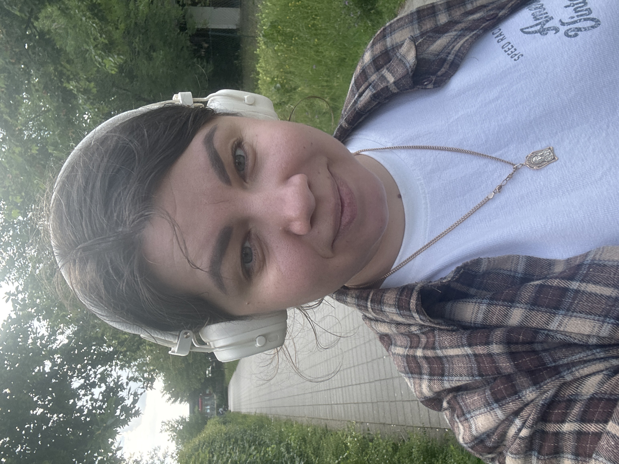 This is a selfie of a young woman wearing white over-ear headphones and a plaid shirt over a white t-shirt. She has dark hair, dark eyebrows, and is smiling at the camera. A gold chain with a pendant is visible around her neck. The background suggests an outdoor setting, with greenery and a paved walkway. The image is taken in daylight, likely during the day, with natural lighting. The woman appears relaxed and happy. No text is visible on the plaid shirt or headphones. The location context is Höhenkirchen-Siegertsbrunn, Germany.