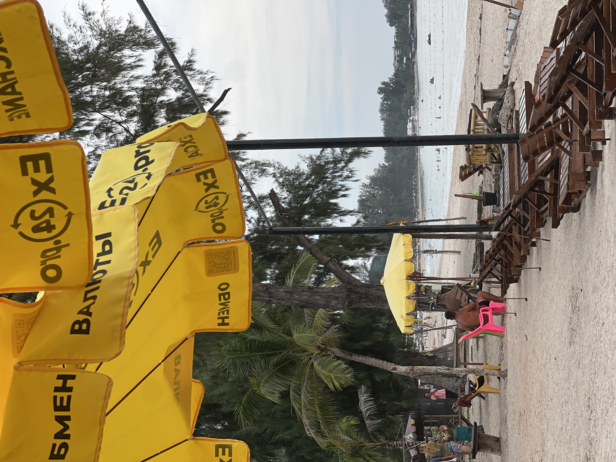 The image captures a scene at a beach in an unspecified location. Rows of yellow umbrellas bearing exchange rate advertisements in what appears to be Russian are visible on the left. To the right, beach chairs are stacked on the sand. Further down the beach, an individual sits on a stack of plastic chairs. The beach stretches to the horizon, where a coastline is visible. The sky is overcast.