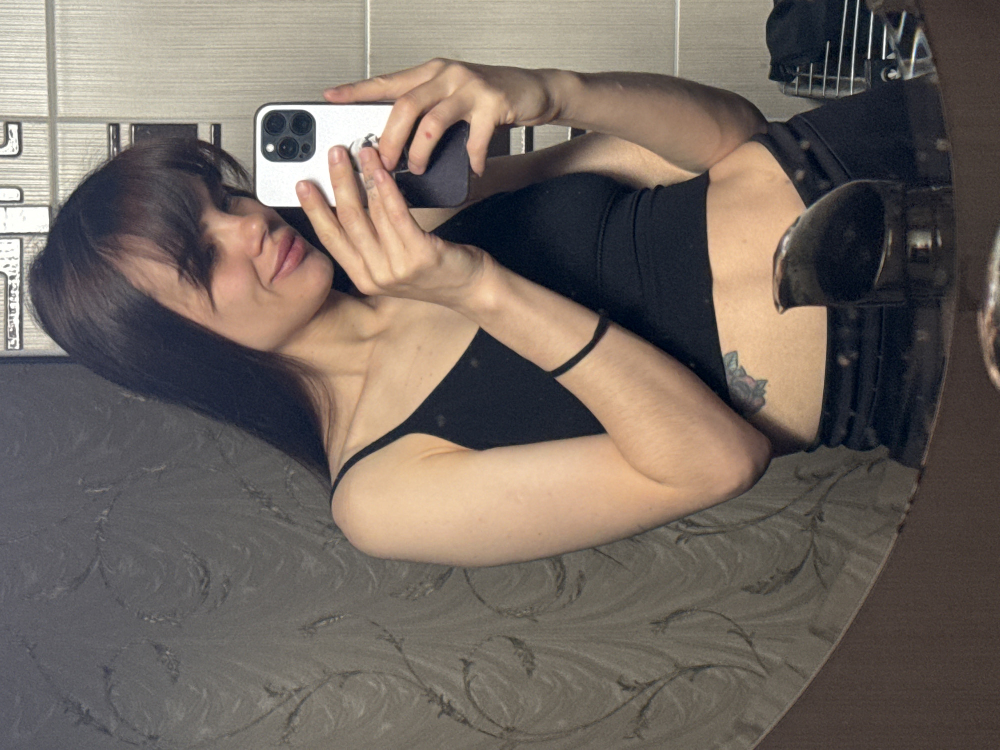 The media file is a mirror selfie featuring a young woman in an indoor setting, likely in Rostov-on-Don, Russia. The image is oriented horizontally, showing the woman as if lying on her side.
**Subjects:** The primary subject is a young woman with dark, shoulder-length hair and bangs. She is wearing a black spaghetti-strap top and what appears to be black bottoms. She holds a white smartphone with a visible Apple logo on its case in both hands, pointing it towards the mirror to capture her reflection. Her left wrist has a black hair tie or bracelet. A floral tattoo, possibly a rose, is visible on her left lower torso/hip area.
**Scene and Setting:** The woman is reclining on a surface covered with a greyish-brown fabric featuring a subtle, swirling floral or leaf pattern. In the reflection behind her, a light-colored tiled wall is visible, suggesting a bathroom or domestic setting. To her reflected left, a dark metal object, possibly a grate or shelving unit, can be seen. The mirror's dark frame borders the right edge of the image.
**Activity:** The woman is actively taking a selfie in a mirror. Her head is slightly tilted, and she has a gentle, closed-mouth smile, indicating a relaxed demeanor.
**Notable Details:** The lighting suggests an indoor environment. Her expression conveys a calm and pleasant mood. No explicit text is visible, apart from the Apple logo on the phone case.