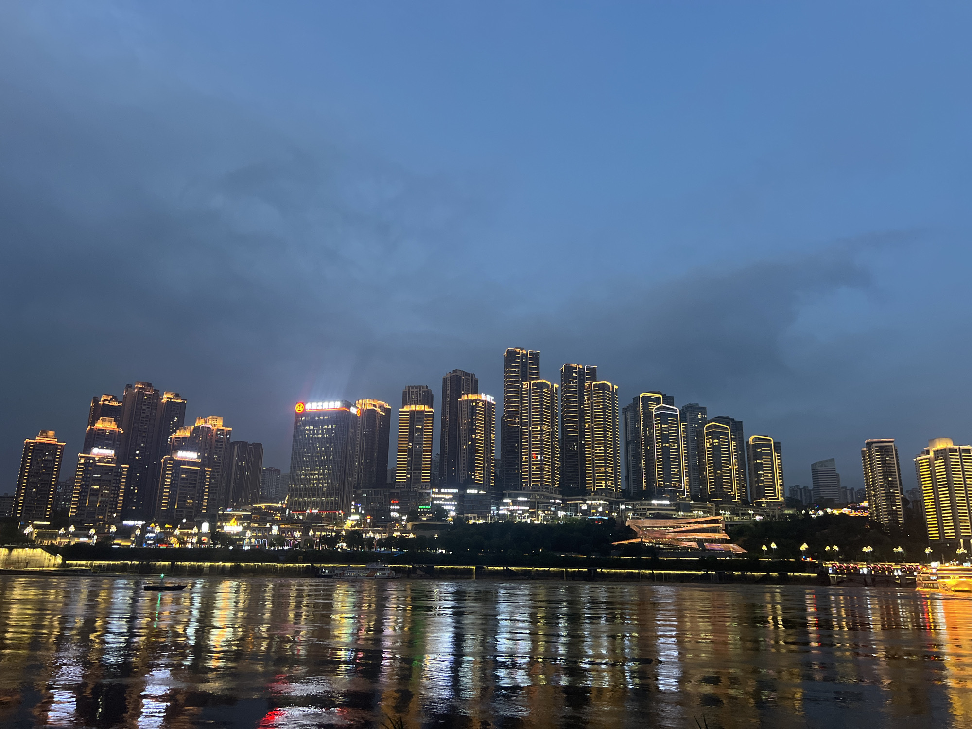 The image depicts a nighttime cityscape along a river, likely in the Jiangbeicheng subdistrict of Chongqing, China, known for its dramatic riverfront development. A dense cluster of modern high-rise buildings with illuminated windows forms the backdrop, their golden light reflecting vibrantly on the dark, rippling water of the river in the foreground. The sky is a deep, dusky blue, transitioning from twilight to night, with subtle cloud formations visible. Along the riverbank, a series of illuminated structures, possibly restaurants or commercial buildings, are visible, their lights creating a continuous luminous band. Several boats are present on the water; one small, dark vessel with a single light sails across the middle ground, while larger, illuminated tourist boats appear further in the distance, their lights contributing to the overall glow on the water. The overall impression is one of a bustling, modern urban environment at dusk, with a focus on architectural illumination and its reflection on the water.