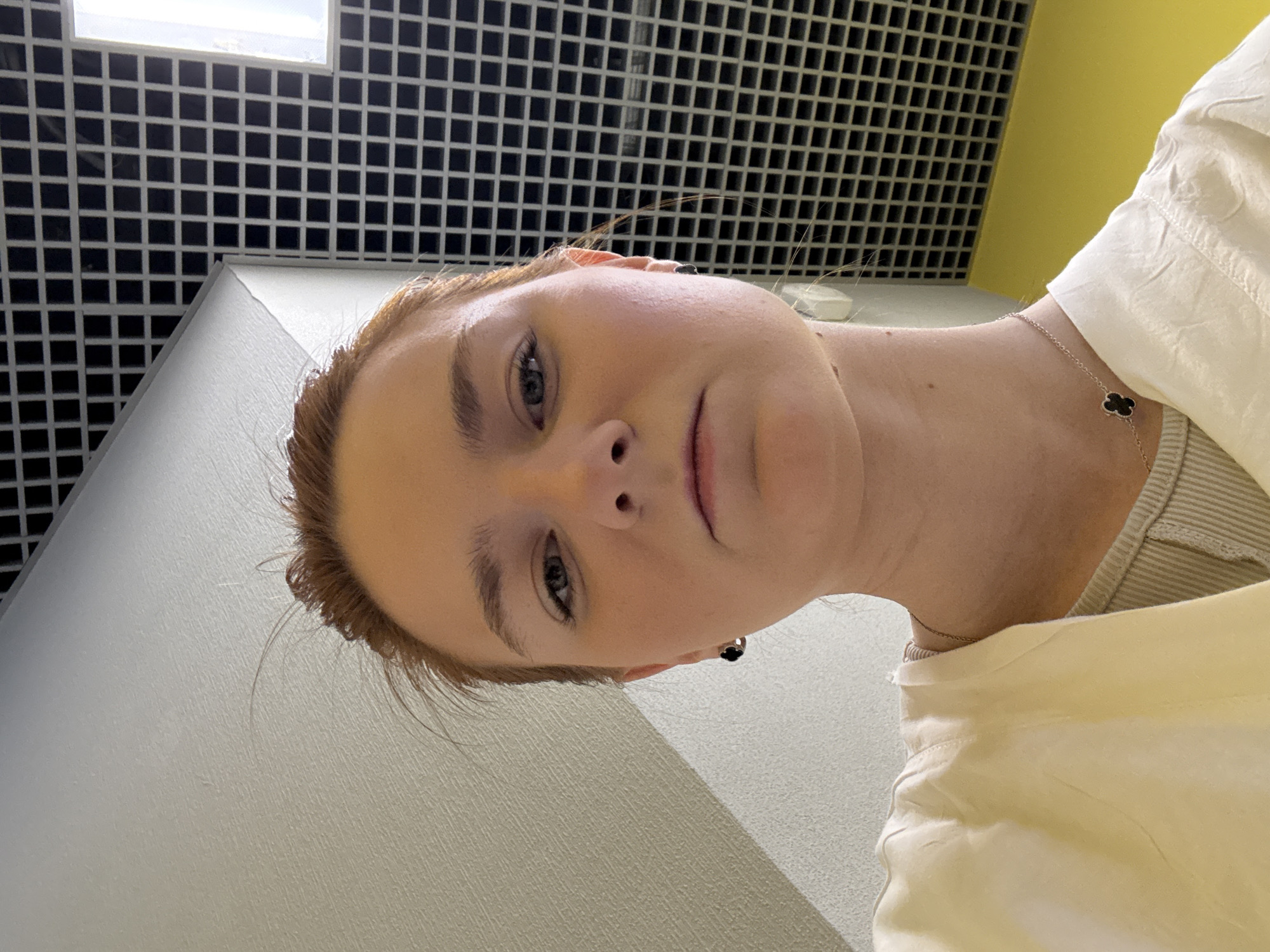 A close-up, low-angle perspective captures a person with fair skin, light brown/reddish hair, and light-colored eyes. The person is wearing light-colored clothing. A delicate necklace with a black clover-shaped pendant is visible on their neck. The person is wearing a black earring.
The background consists of a grid-like ceiling, a neutral-colored wall, and a yellow wall.
The location is in the city of Unknown, Russia.