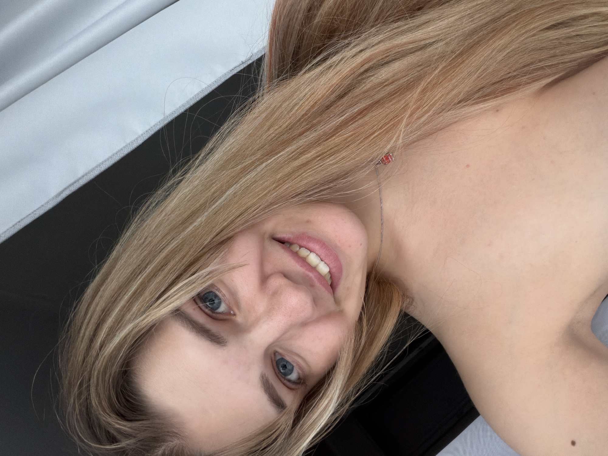 A blonde-haired woman with blue eyes and a fair complexion is angled in the frame, smiling slightly at the viewer. She is wearing a delicate silver necklace with a small red pendant. The background shows a mix of white and black surfaces. Location details are not discernible from the image provided.