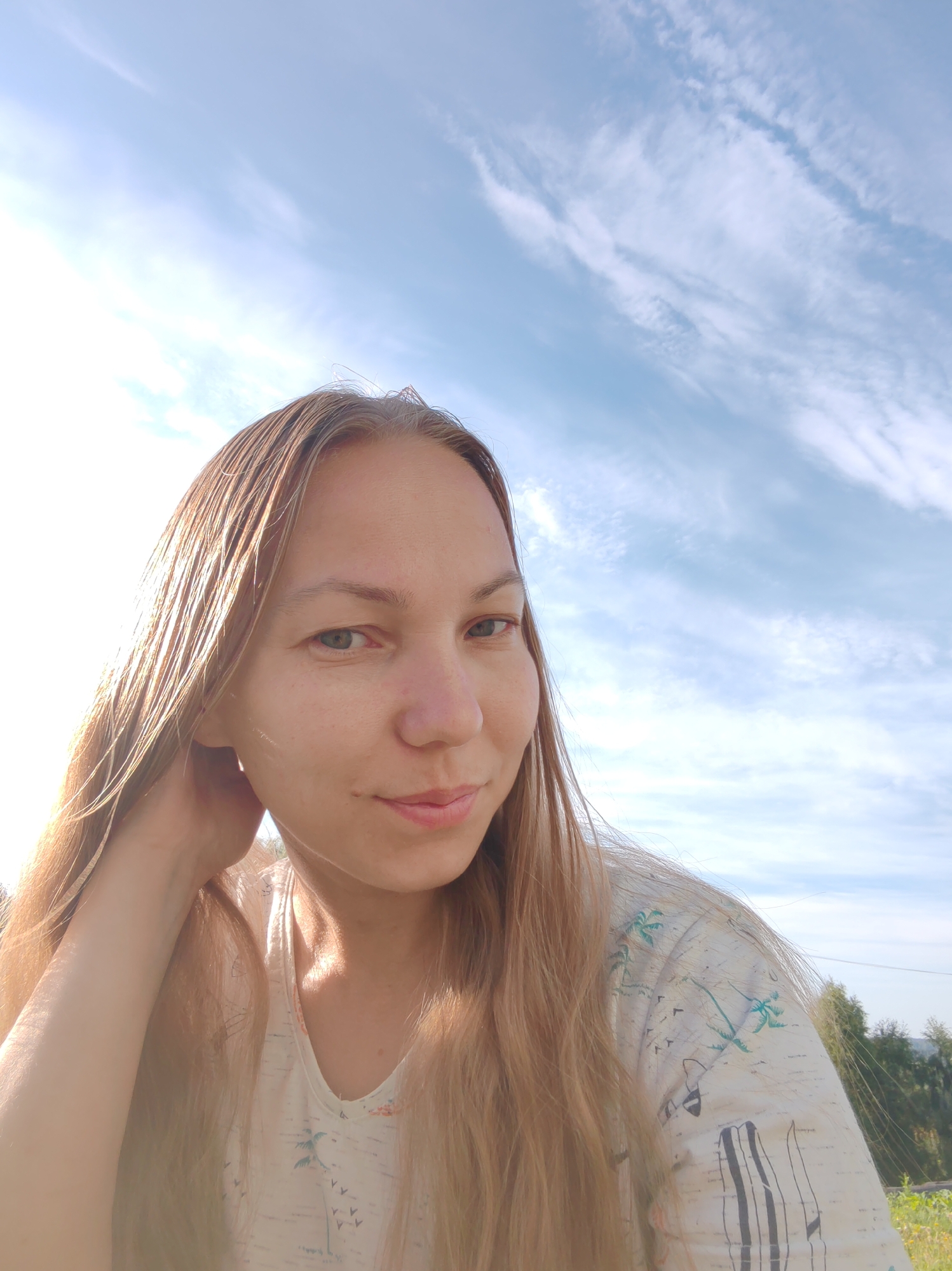 A woman with long, blonde hair is taking a selfie outdoors. She is wearing a light-colored t-shirt with a pattern of palm trees and birds. The background is a bright blue sky with wispy white clouds and a hint of greenery from trees and vegetation. The lighting suggests it is daytime, possibly late afternoon, due to the soft, warm glow. The scene appears to be in a rural or suburban area, possibly on the outskirts of the city of Chukhloma, Russia, given the natural light and open sky.