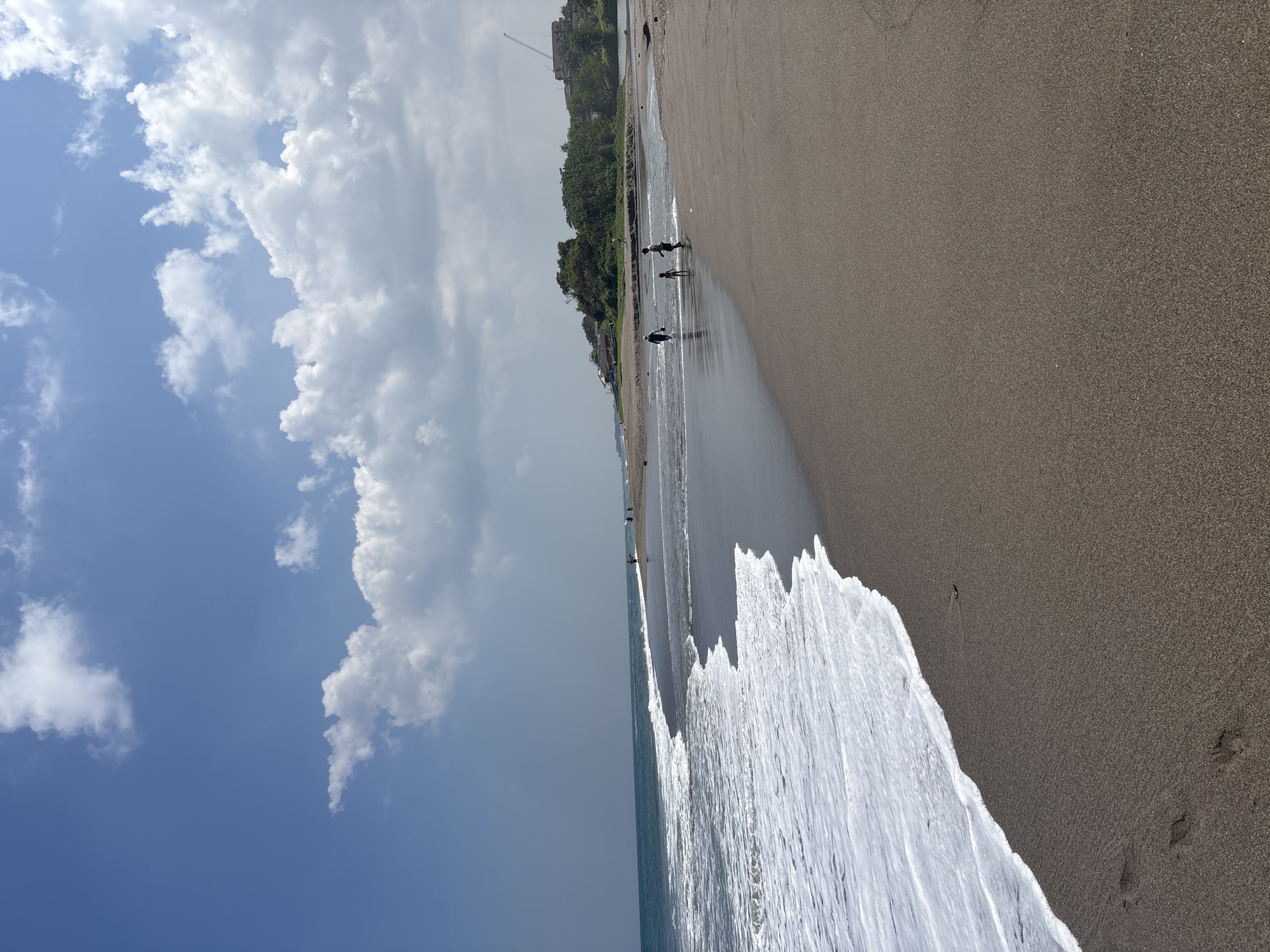 The image depicts a wide, dark sand beach in North Kuta, Indonesia, with the ocean on the left and land on the right. Gentle waves with white foam are rolling onto the shore. In the distance, a few people are visible walking along the beach. The land beyond the beach is covered with lush green trees and some buildings, including a construction crane in the background. The sky is blue with scattered white clouds. The overall scene conveys a sense of a serene coastal environment.