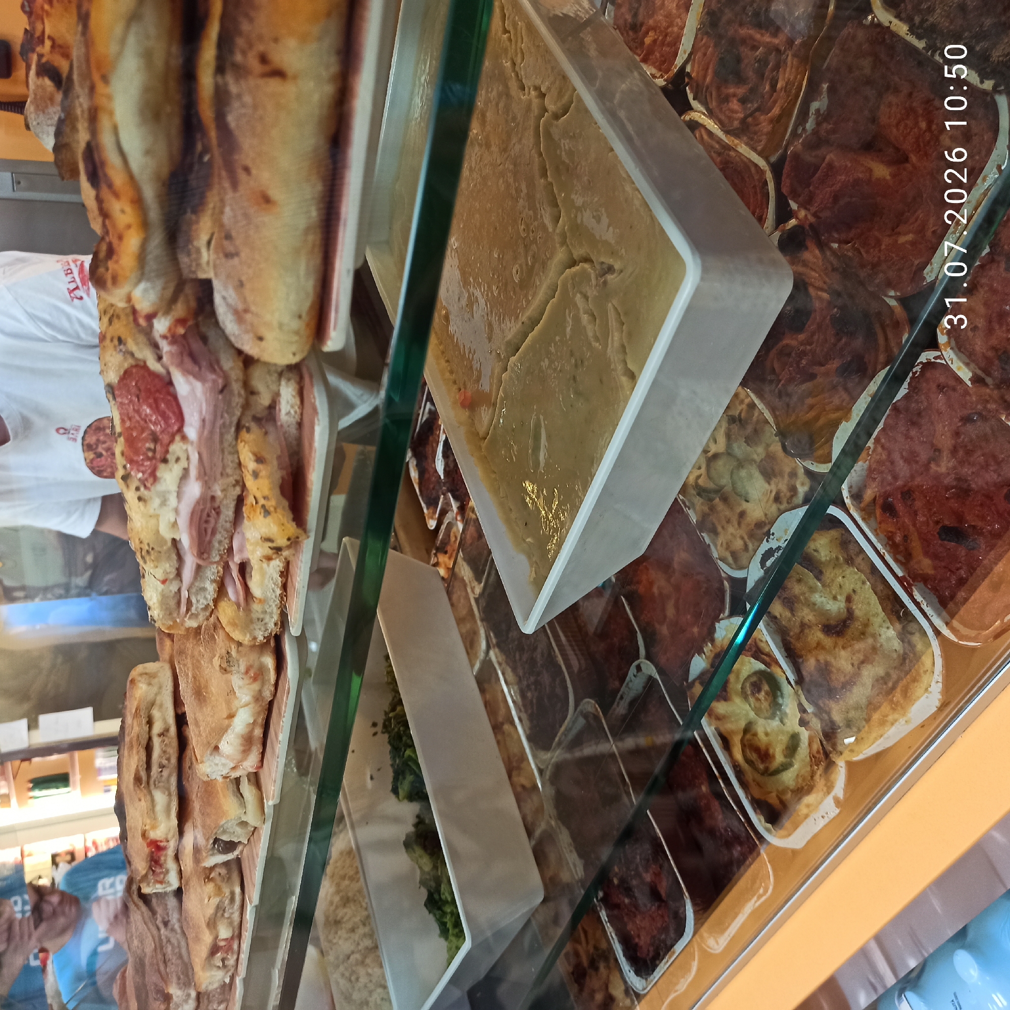 This is a close-up, eye-level shot of a food display case filled with various prepared foods, predominantly savory pastries and what appear to be casseroles or baked dishes. The display is lit from above, creating reflections on the glass surface.
In the foreground, a white rectangular tray holds a pale, creamy, semi-liquid substance, possibly a purée or a thick sauce, which shows some cracking on its surface. To the right and below this tray, several smaller aluminum foil pans contain baked goods with golden-brown crusts, some with visible fillings like red sauce and vegetables.
Further back, in a deeper white rectangular tray, is a pile of cooked green vegetables, possibly broccoli or spinach. Behind that, several larger, golden-brown pastries, some folded or stuffed, are stacked and presented on white and red patterned plates. One large pastry in the center is a sandwich-like creation with layers of thinly sliced pink meat (likely ham or prosciutto) and possibly cheese, topped with sliced red tomatoes.
In the background, a person wearing a blue t-shirt and glasses is partially visible, leaning over the display. They are looking down at the food. Another person in a white t-shirt with a logo is also partially visible behind the food display. The environment appears to be a food establishment, possibly a bakery or deli, with shelves and products visible in the blurred background.
The timestamp "31.07.2026 10:50" is overlaid on the bottom right of the image. The presence of "BARI" on the white t-shirt suggests a possible connection to the city of Bari, which is in the vicinity of Modugno, Italy. However, there are no definitive visual cues within the image itself that confirm the specific location as Modugno, Italy.