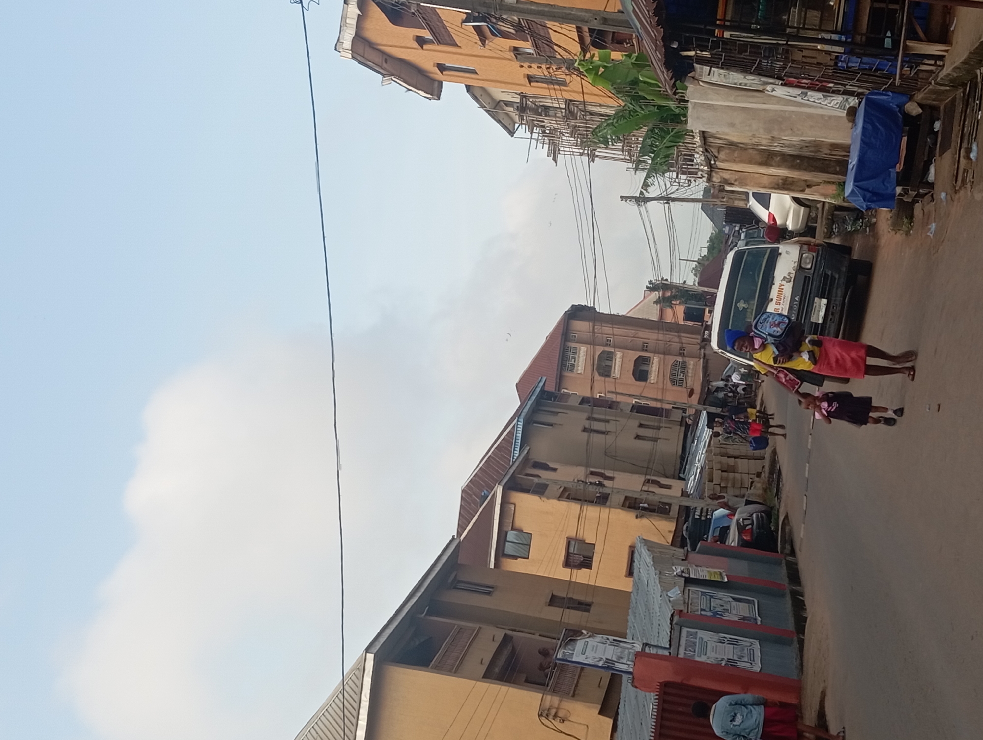 The image depicts a street scene in Nkpor, Nigeria. The street is lined with multi-story buildings, some of which appear to be residential. Overhead, there are power lines. On the right, a white van is parked, and several people, including children in school uniforms, are walking along the road. There are also various objects along the street, such as metal structures and what appear to be advertisements posted on walls. The sky is overcast.