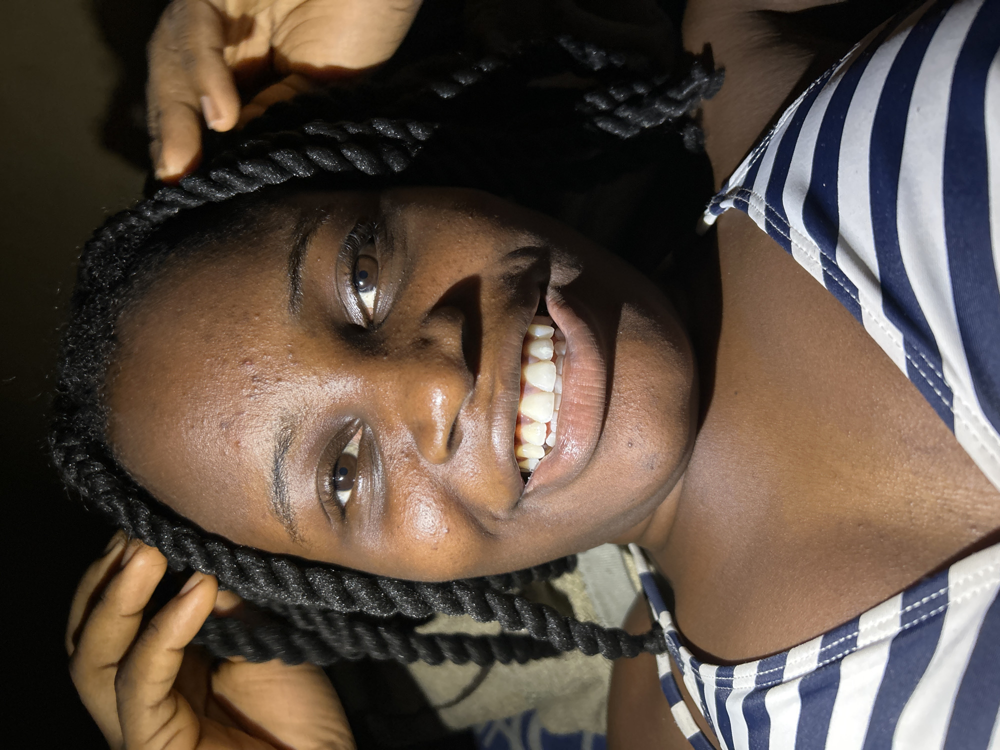 A close-up, head-and-shoulders shot shows a young Black woman with dark skin and braided hair smiling broadly. She is wearing a blue and white striped bikini top. Her hands are visible near her head, one on each side, as if adjusting her braids. The lighting is dim, creating shadows on the dark background. The image appears to have been taken in a dimly lit indoor setting, possibly a bedroom, in Ihiagwa, Nigeria.
