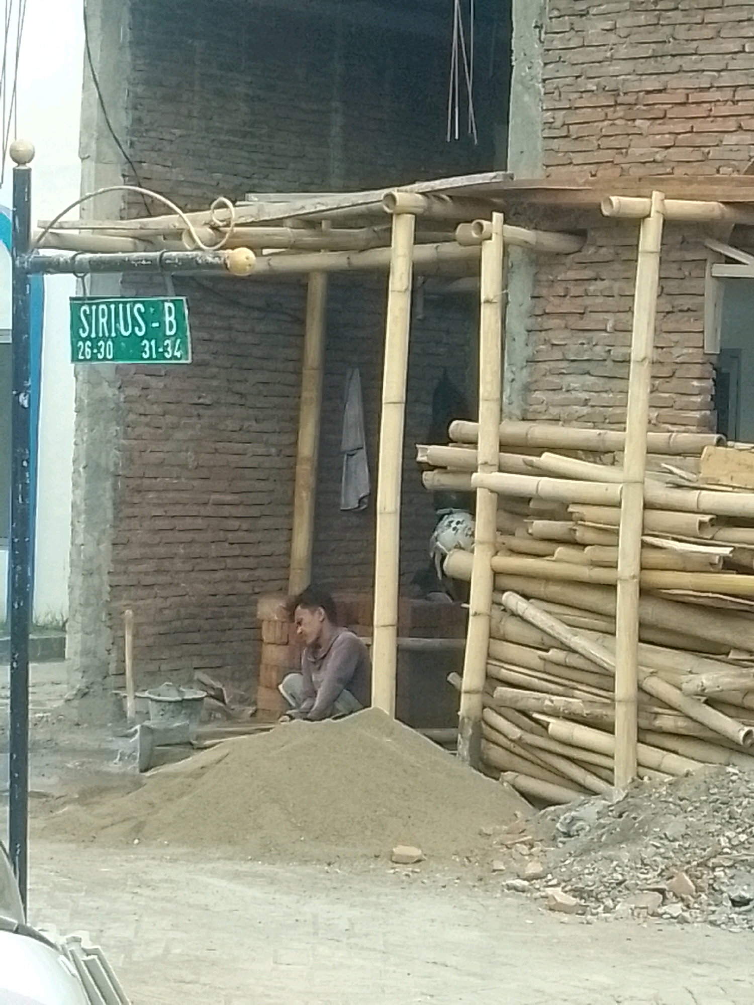 The image depicts a construction site in Jember, Indonesia. A street sign labeled "SIRIUS-B 26-30 31-34" hangs on a pole to the left. In the center, a construction worker, wearing a grey long-sleeved shirt and grey pants, is seated on a pile of sand, resting. The background shows a brick wall with bamboo scaffolding. Piles of sand and construction materials are scattered around the site. The overall scene suggests an active construction project.