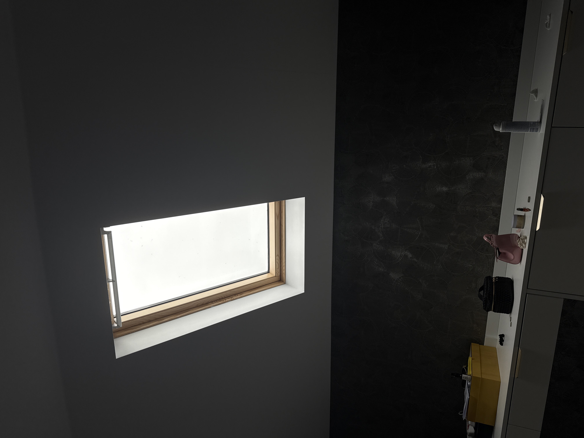 The image shows an interior view. A rectangular window is set into a gray wall, letting in natural light. To the right, a shelf or countertop runs vertically against a darker, textured wall. Various objects rest on the shelf, including a pink purse, a black bag, and a yellow box. The overall composition suggests a room with a skylight and a minimalist design. The image does not contain sufficient information to reliably determine the location as Plovdiv, Bulgaria.
