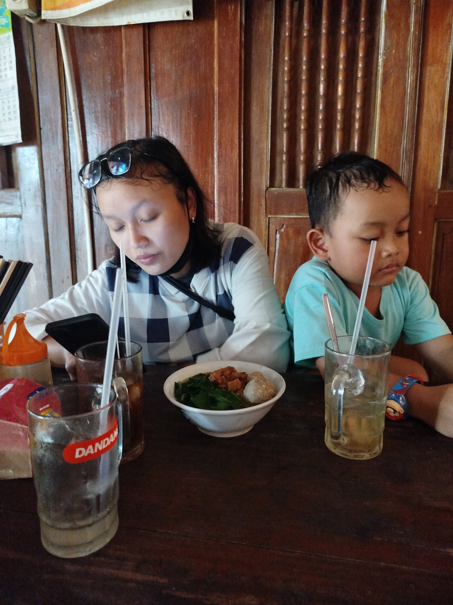 The image depicts two individuals, an adult female and a young male, seated at a wooden table. The female, with glasses perched on her head, is looking down, her face partially obscured by her hair. She is wearing a white and blue checkered long-sleeved shirt and a black neck strap. The young male, wearing a light blue t-shirt, is also looking downwards.
In front of them are two glasses with straws, a bowl of food containing noodles, leafy greens, and meatballs, and a red and orange dispenser. A smartphone is visible on the table near the female. The background consists of a wooden wall with vertical planks. The scene appears to be an indoor setting, possibly a casual dining establishment, in Pojok, Indonesia.