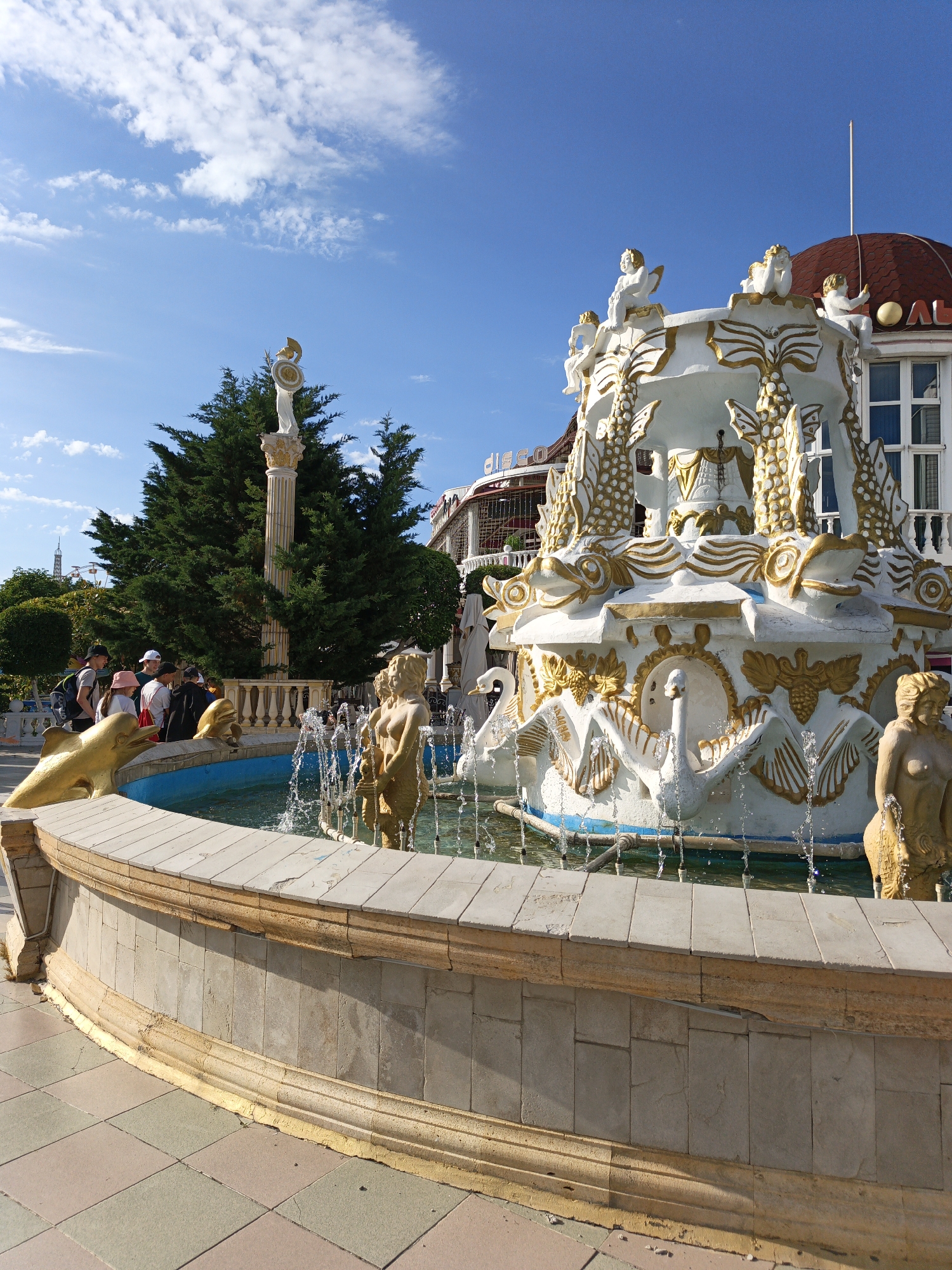 The image captures a vibrant, sunny outdoor scene featuring an ornate fountain in a public area of Kabardinka, Russia.
The central subject is a large, multi-tiered fountain, predominantly white with intricate gold accents. Its base features sculptures of swans and golden mermaid statues, from which water jets spray into the pool. Golden dolphin figures also adorn the edge of the fountain basin. The fountain's main body is decorated with fish-like designs, while the uppermost tier is adorned with several white cherub-like figures.
To the left of the fountain, a group of casually dressed individuals, appearing to be young adults or teenagers, are observing the scene. Behind them, lush green trees frame a tall column topped with a white statue resembling a warrior, holding a golden helmet and shield. In the distant background, a faint outline of a Ferris wheel is visible.
To the right, a building with a distinctive red-domed roof and white window frames can be seen. Partially visible text on the building reads "Disco" and another fragment "ОЛЬГ".
The time of day is bright and sunny, indicated by the clear blue sky with scattered white clouds and strong illumination. The ground is paved with square tiles in alternating shades of light beige/pink and grey/green. The fountain's stone perimeter wall features a golden trim, which shows some signs of wear. The overall atmosphere is lively and typical of a public attraction or resort area.