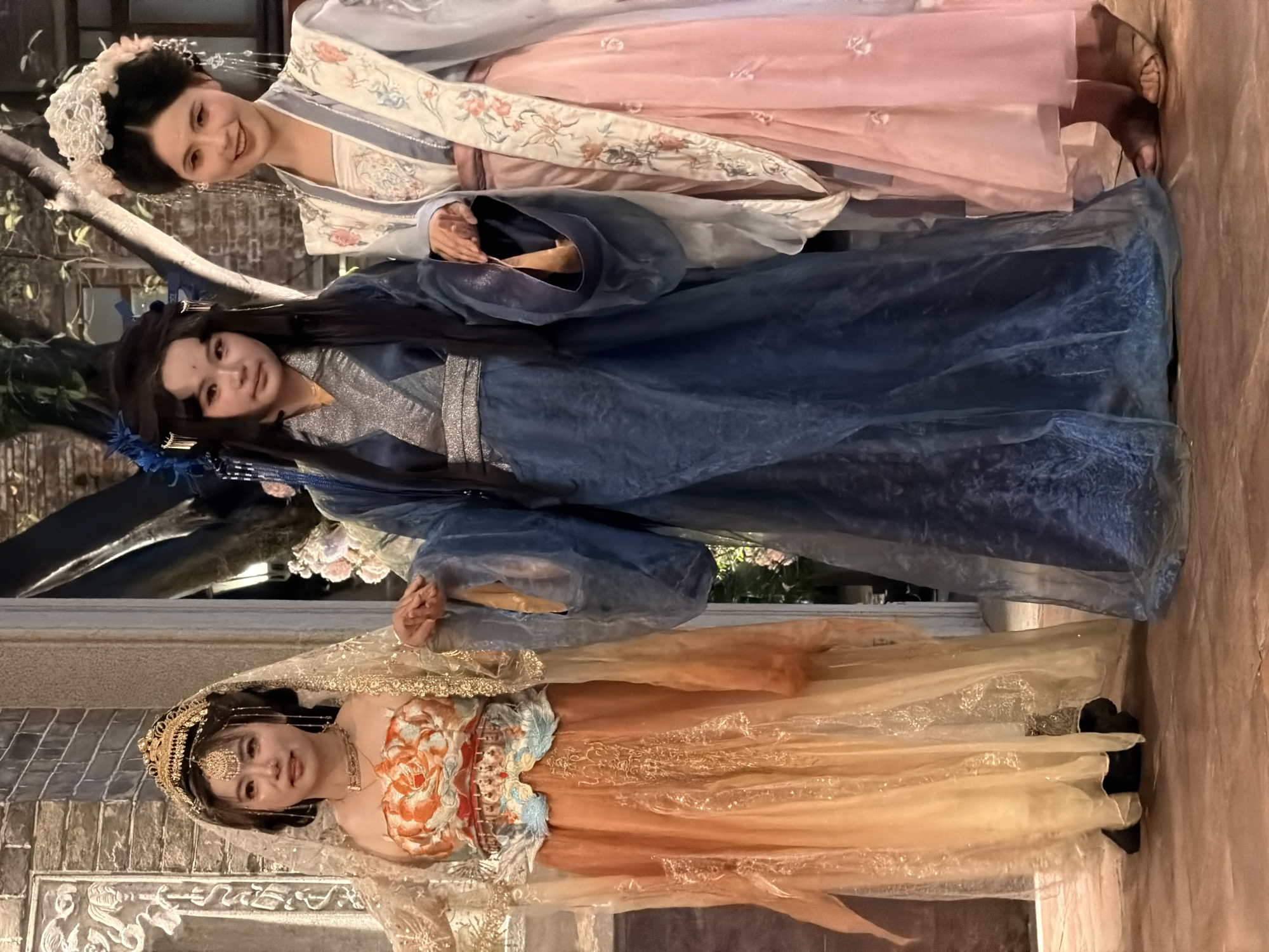 Three young women are posing for a photo in traditional Chinese attire, likely during a cultural event or photoshoot. The woman on the left wears an elaborate golden crown with dangling embellishments and a flowing orange and yellow gown with an embroidered bodice. The woman in the center is dressed in a deep blue, layered robe with wide sleeves and a silver sash. The woman on the right is adorned in a light pink skirt and a white and blue patterned outer garment, with a white floral headdress. They are standing on a stone-like floor with a brick wall and foliage in the background, suggesting an outdoor setting or a carefully constructed set designed to evoke a historical ambiance. The lighting appears to be artificial, creating a warm glow on the subjects.