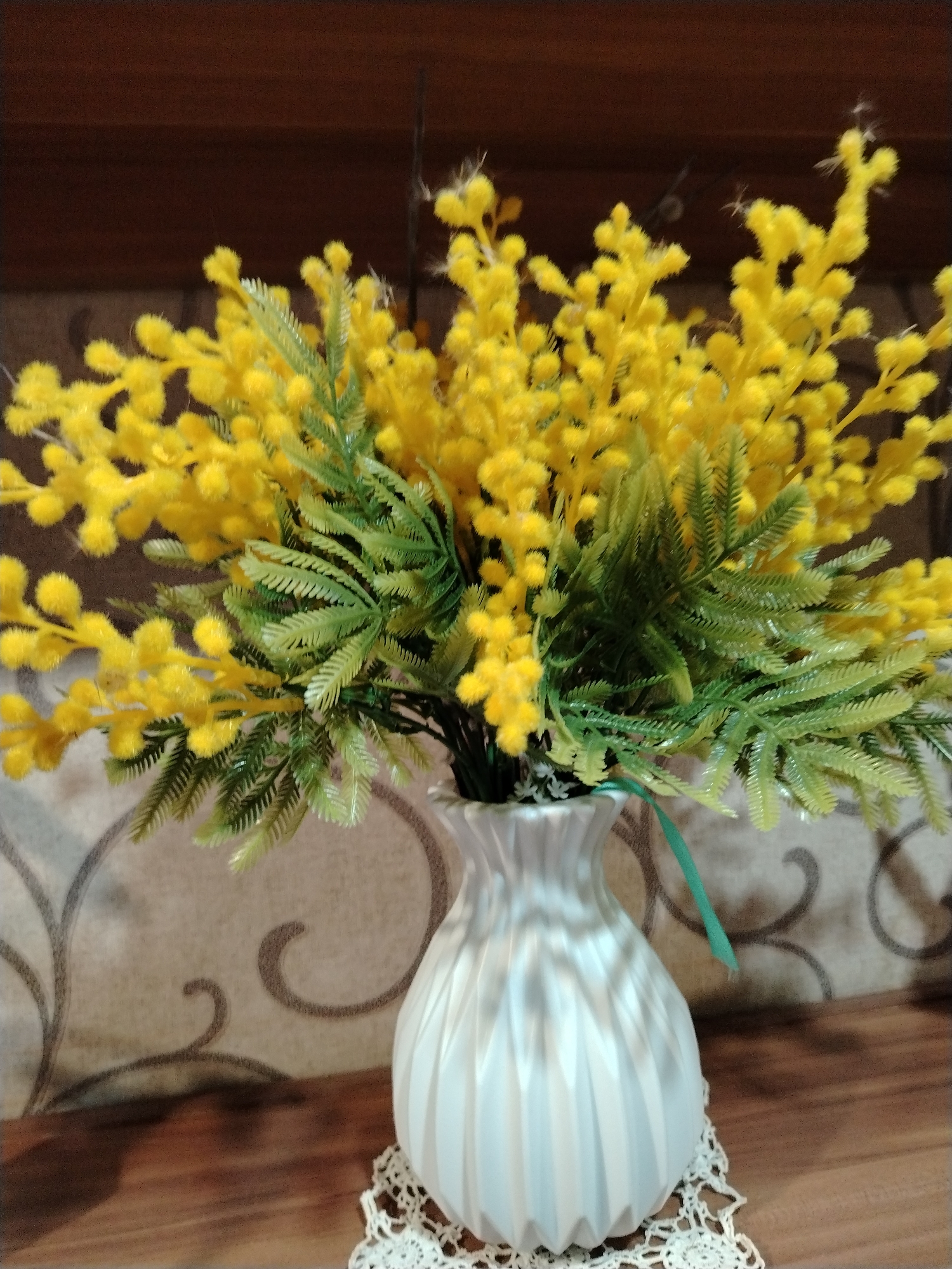 The image presents an indoor still life, prominently featuring a bouquet of artificial flowers in a white vase. The bouquet consists of numerous bright yellow, fluffy blossoms, strongly resembling mimosa, interspersed with vibrant green, artificial foliage that has a distinct plastic texture. A thin green ribbon or string is visible hanging from within the bouquet, draping slightly over the vase's side.
The vase itself is white with a textured, ribbed, or fluted surface that creates vertical lines and light reflections. It is placed on a delicate, white, circular crocheted doily, which sits on a warm, medium-brown wooden surface, likely a table or shelf.
In the background, a wall is visible, covered with light-colored wallpaper featuring a repeating pattern of subtle, dark grey, swirling or scrolling designs. Above the wallpaper, a darker, horizontal brown wooden panel or shelf extends across the upper portion of the frame.
The lighting in the image appears to be soft and indirect, illuminating the arrangement without harsh shadows, suggesting an indoor setting with ambient light, possibly daytime or well-lit artificial indoor light. There are no people, visible text, or discernible activities taking place. The scene depicts a decorative arrangement within what appears to be a home environment in Zhukovo, Russia.