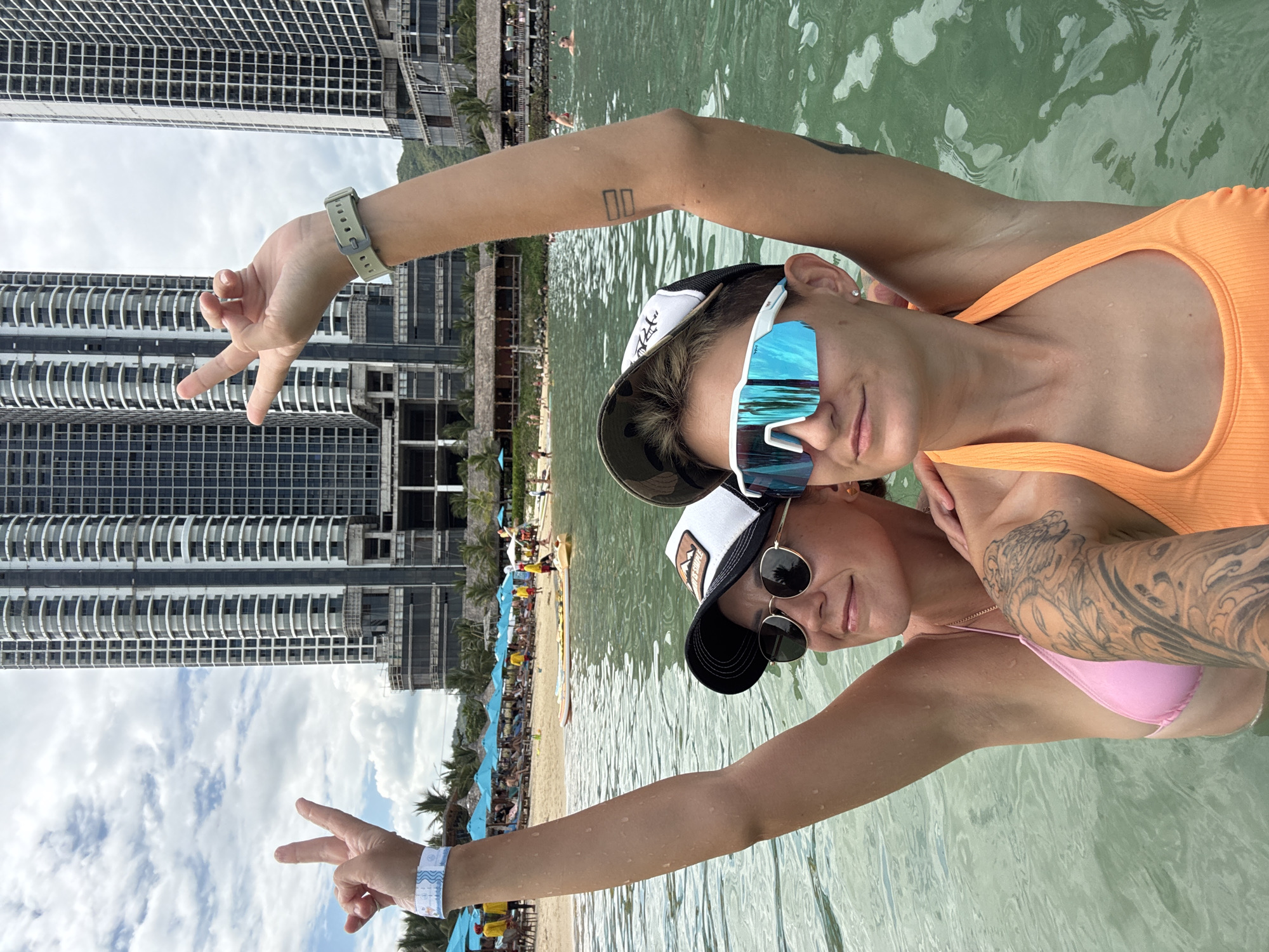 Two women are posing for a selfie in the ocean in front of tall buildings in Nha Trang, Vietnam. The woman on the left is wearing a pink bikini top and sunglasses. She has her right arm raised and is making a peace sign. The woman on the right is wearing an orange bikini top and futuristic sunglasses. She has her left arm raised and is also making a peace sign. The water is clear and green, and there are some buildings and umbrellas visible on the beach in the background. The sky is cloudy.