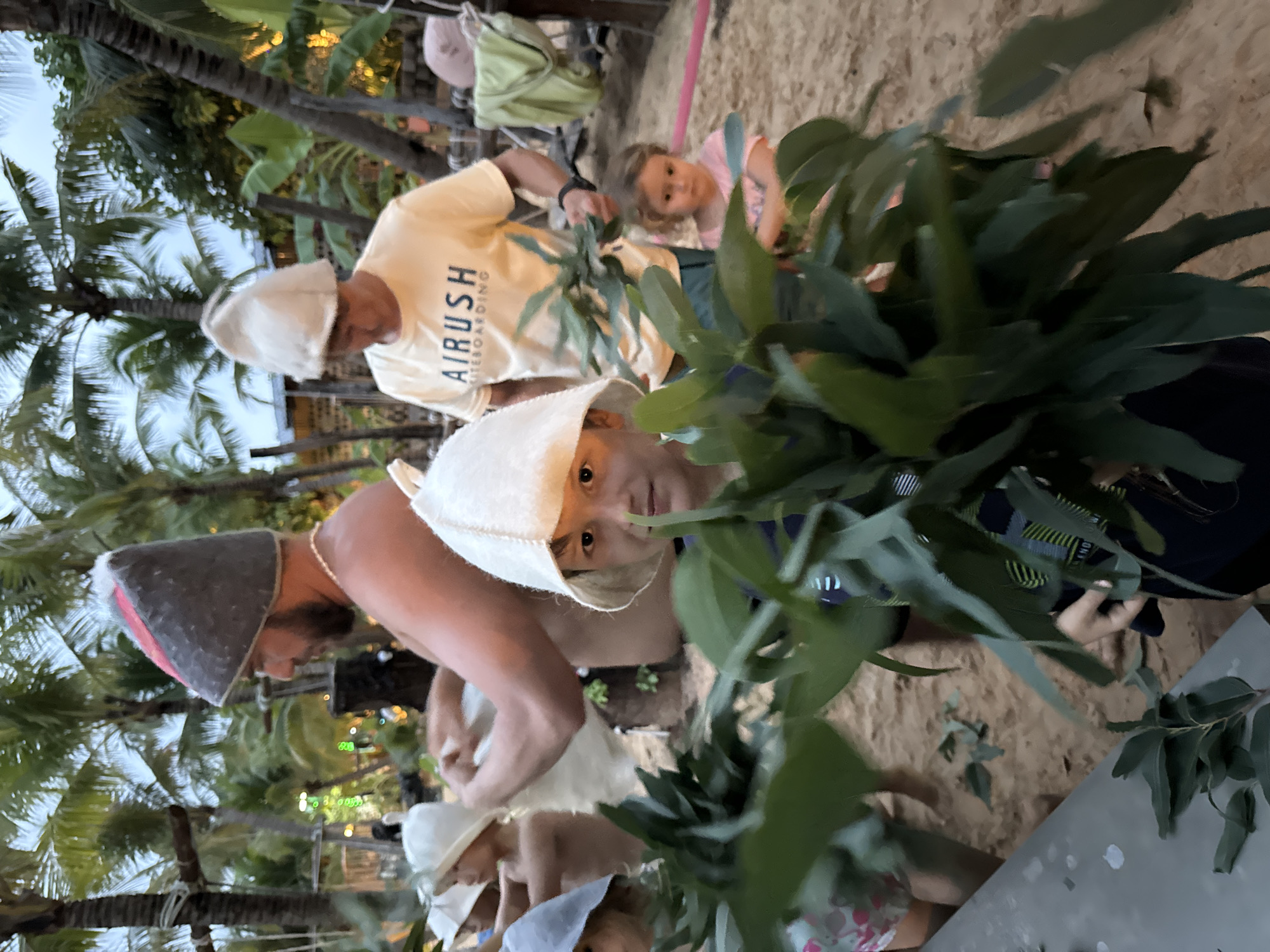 In Mũi Né, Vietnam, the scene captures a group of people on a sandy surface outdoors. One person wears a light-colored shirt with "AIRUSH KITEBOARDING" printed on it and a white hat. There are several other individuals nearby, including a child looking directly at the camera, partially obscured by a large bouquet of green leaves. A man with a beard is wearing a gray and red hat and is partly visible. Palm trees and other vegetation are visible in the background, suggesting a tropical or coastal environment. The scene appears candid, possibly taken during a casual moment.
