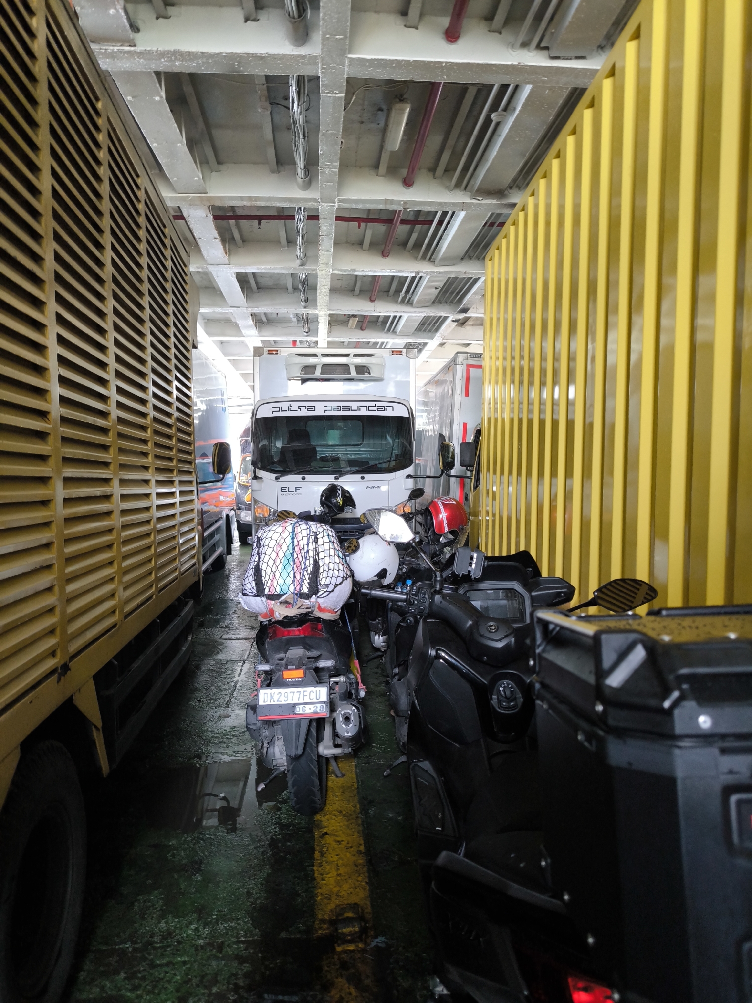 The image provides a low-angle view of a densely packed vehicle deck, likely on a ferry or cargo ship. Vehicles are lined up in a narrow passage, suggesting they are either in transit or waiting to be unloaded.
In the foreground to the left, the side of a large yellow truck or cargo container with horizontal slat-like paneling is prominent, with its tire partially visible. Directly opposite, on the far right, a similar yellow structure with vertical slats acts as a wall or barrier.
In the center of the frame, a white box truck faces away from the camera. The words "putra pasundan" and "ELF" are visible on its front grille and windshield, respectively. Parked closely around this truck and along the passage are several motorcycles and scooters. One scooter in the immediate foreground, positioned centrally, carries luggage secured with a colorful net on its rear seat. Its license plate clearly displays "DK 2977 FCU" and "06-28". To the right, a larger, dark-colored motorcycle or maxi-scooter is visible, equipped with a black, hard-shell storage box on its rear. A red helmet rests on the handlebars of one of the motorcycles.
The floor of the deck is visibly wet and appears somewhat grimy, reflecting the ambient light. Above, the ceiling reveals an industrial structure of exposed beams, supports, electrical conduits, and red pipes, along with visible lighting fixtures. No people are discernible in the scene. The overall impression is one of organized transport within a functional, robust environment.