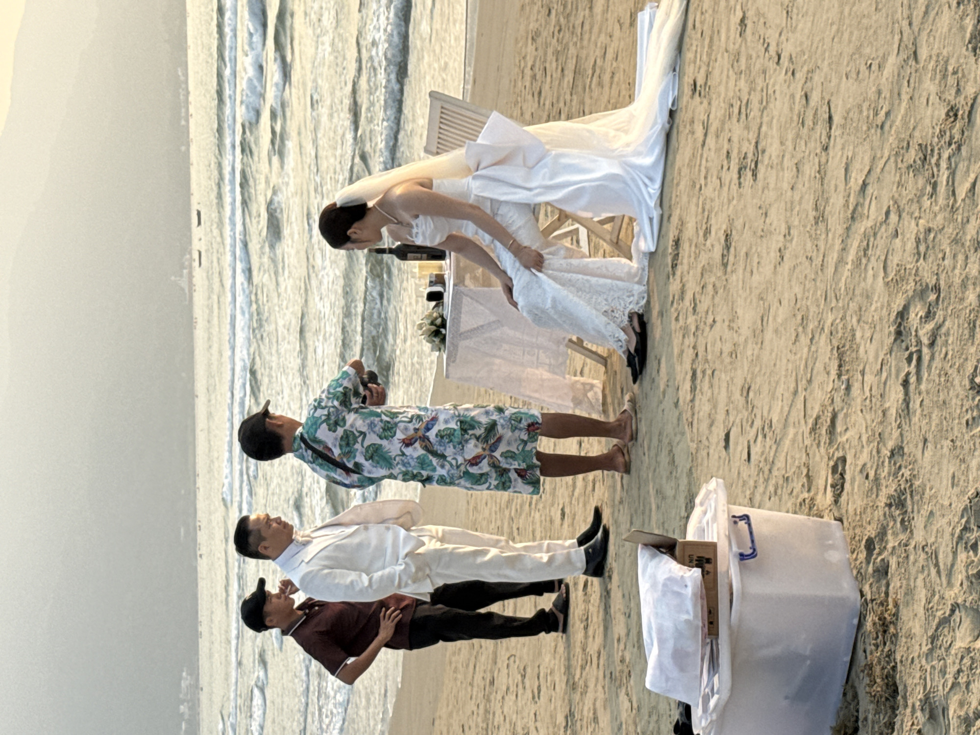 A daytime outdoor scene depicts a wedding ceremony taking place on a beach. The location is in Đà Nẵng, Vietnam, as evidenced by the coastal environment and the presence of Asian individuals.
The bride, dressed in a long white gown and veil, stands next to a white table draped with a white cloth. A bottle of wine, flowers, and candles are on the table. She is interacting with her dress.
Three men stand to the side, one wearing a white suit and two dressed casually. There is a white bin next to them.
The beach setting includes sand and the ocean with gentle waves. The sky is overcast.