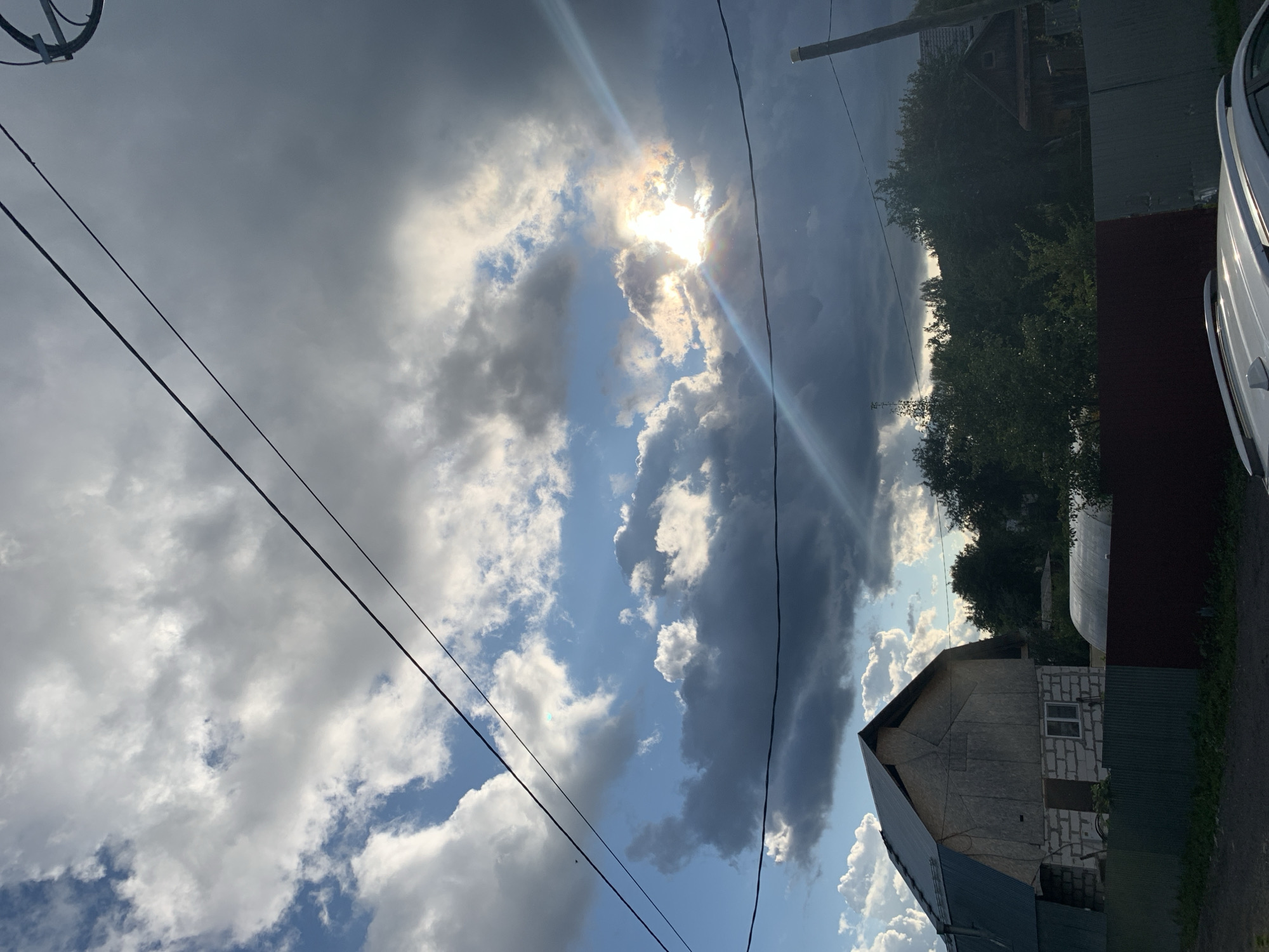 In Asha, Russia, this image captures a late afternoon sky filled with dramatic clouds. The sun breaks through in the upper right quadrant, casting a bright glare and a light flare across the scene. Below the sky, a partially constructed building with a metal roof stands on the left, constructed from white bricks. A green metal fence runs along the foreground, with a section of dark red fencing to its right. Beyond the building and fence, dense green trees create a dark backdrop. In the far right of the image, a wooden structure and a utility pole are visible. Power lines traverse the sky from the left to the right edge of the frame. The overall scene suggests a rural or suburban setting with an overcast sky, typical of the region.