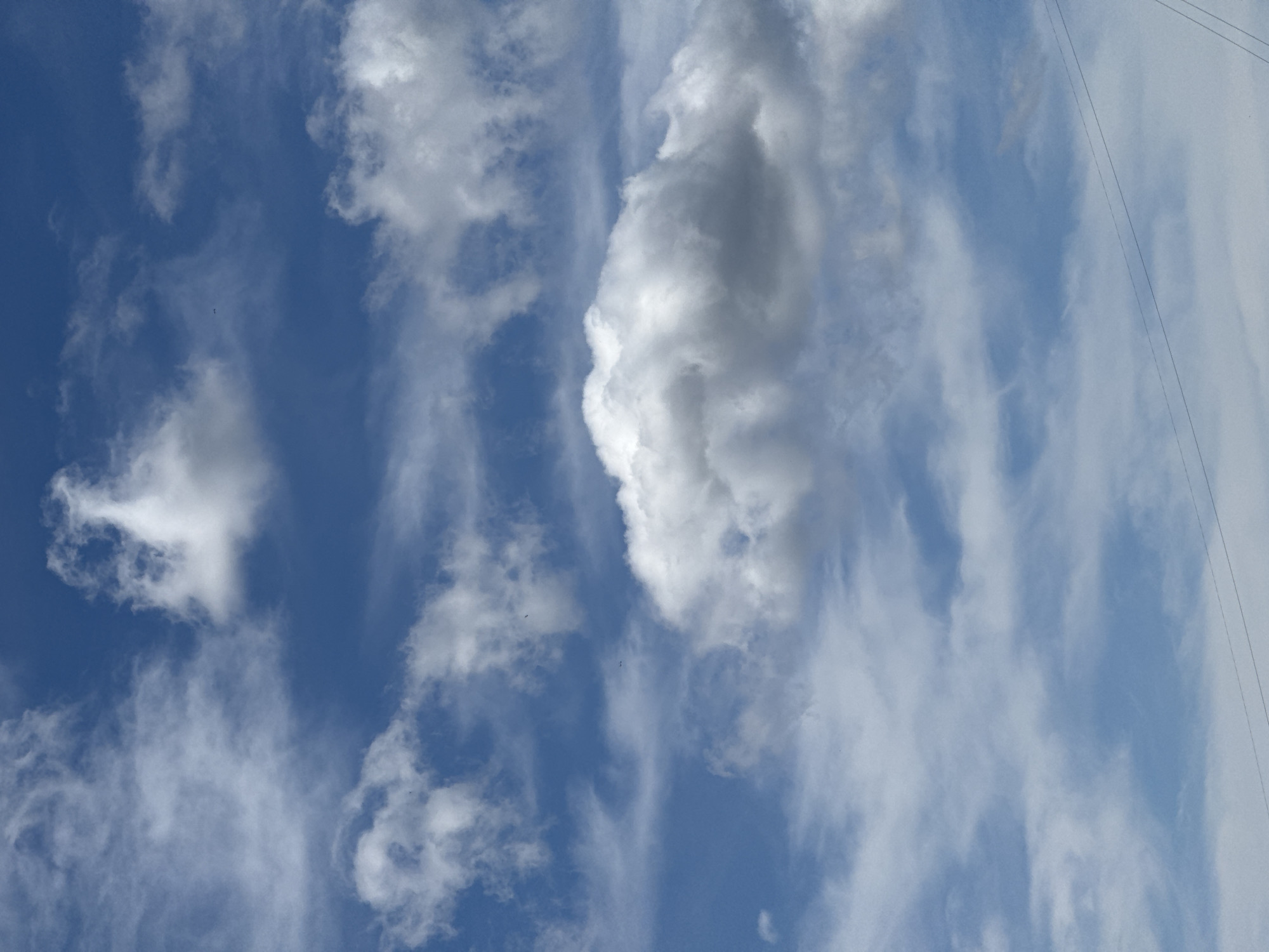 The image shows a bright blue sky with scattered white clouds. There are two thin, dark, horizontal lines visible in the lower portion of the image, which appear to be power lines. The clouds vary in density and shape, with some appearing wispy and others more solid and billowy. The overall scene is serene and depicts a clear day. There are no people, buildings, or any other identifiable landmarks visible in the image. The image does not provide any specific information or visual cues that would indicate its location within the city of Krasnoyarsk, Russia.