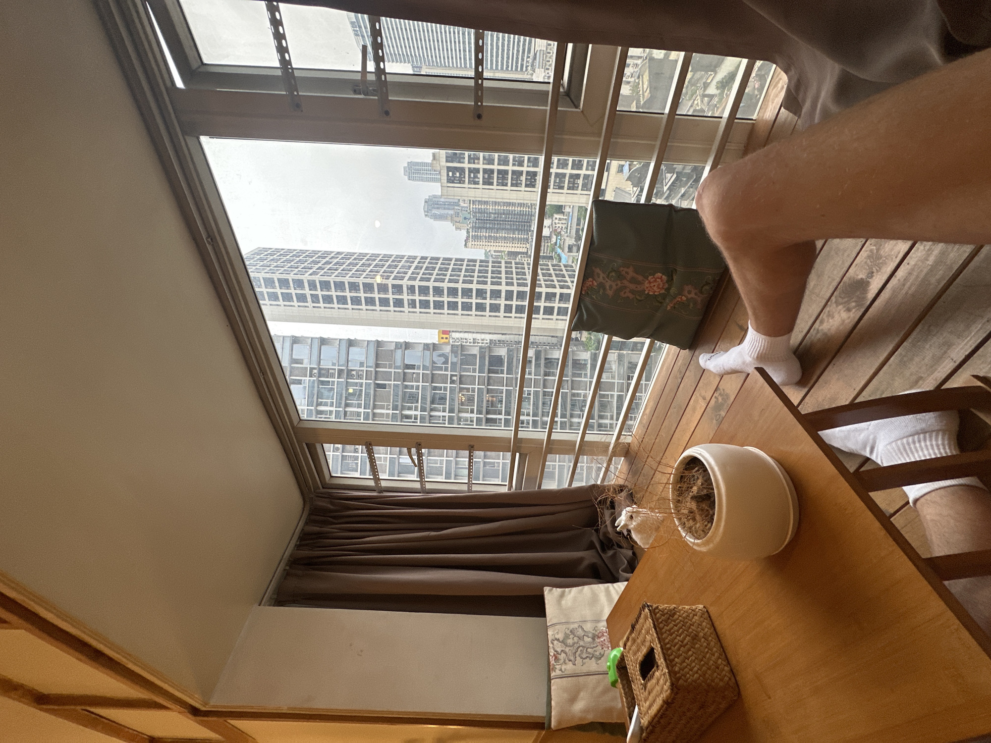 A high-angle, indoor shot depicts a room with a wooden table bearing a potted plant and a woven basket. A partial view of a person's legs in white socks is visible. The room has a large window overlooking a cityscape in Yuzhong District, China, with numerous high-rise buildings under a cloudy sky. Brown curtains partially cover the window.