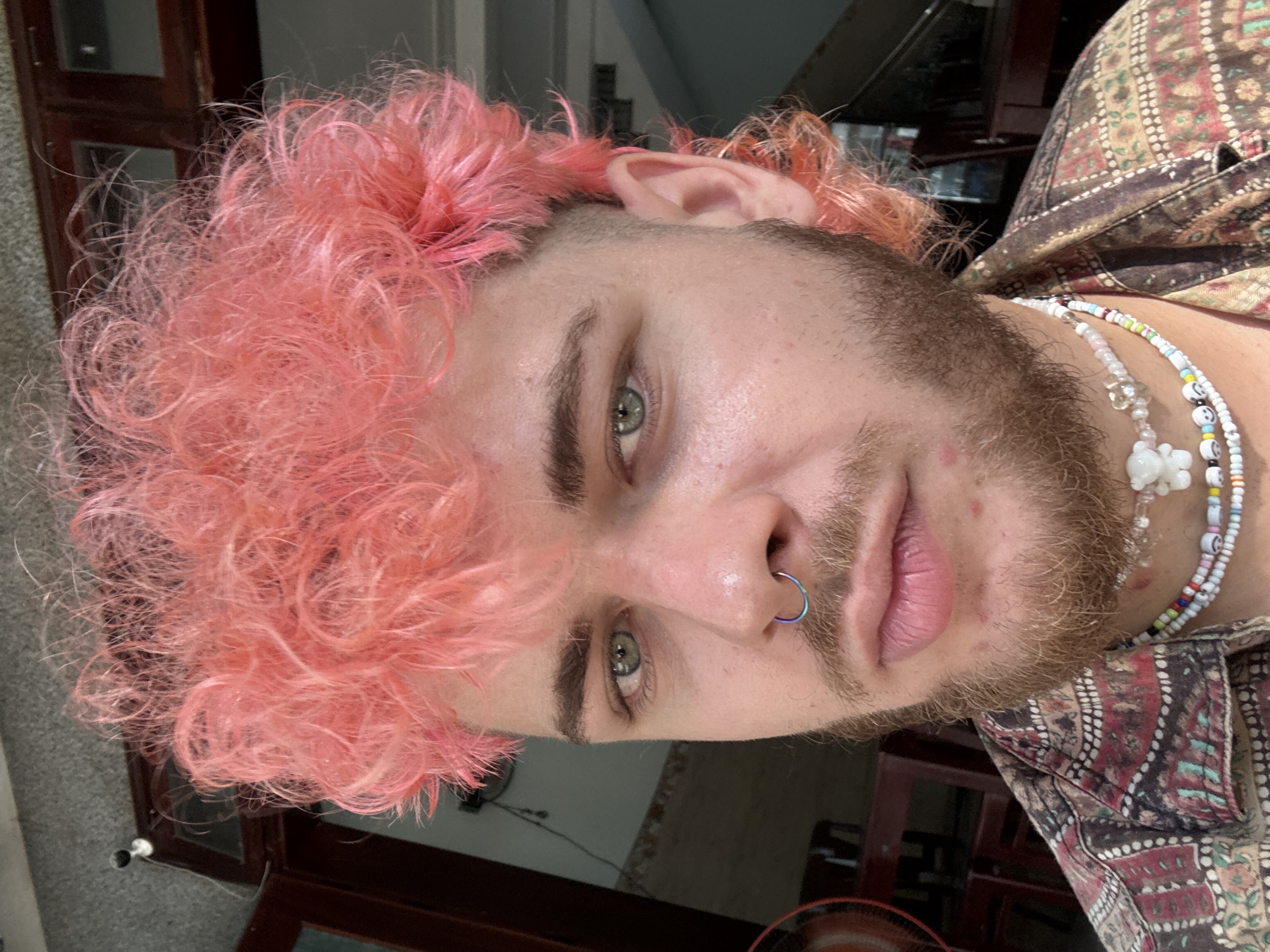 A close-up, slightly rotated portrait of a person with pink curly hair, a nose ring, a reddish-brown beard, and fair skin. The person has light green eyes and is wearing a patterned shirt. A beaded necklace is visible. The background suggests an interior setting, with a window and door frame partially visible. The location is not discernible to be Đà Nẵng, Vietnam, based on the information in the image.