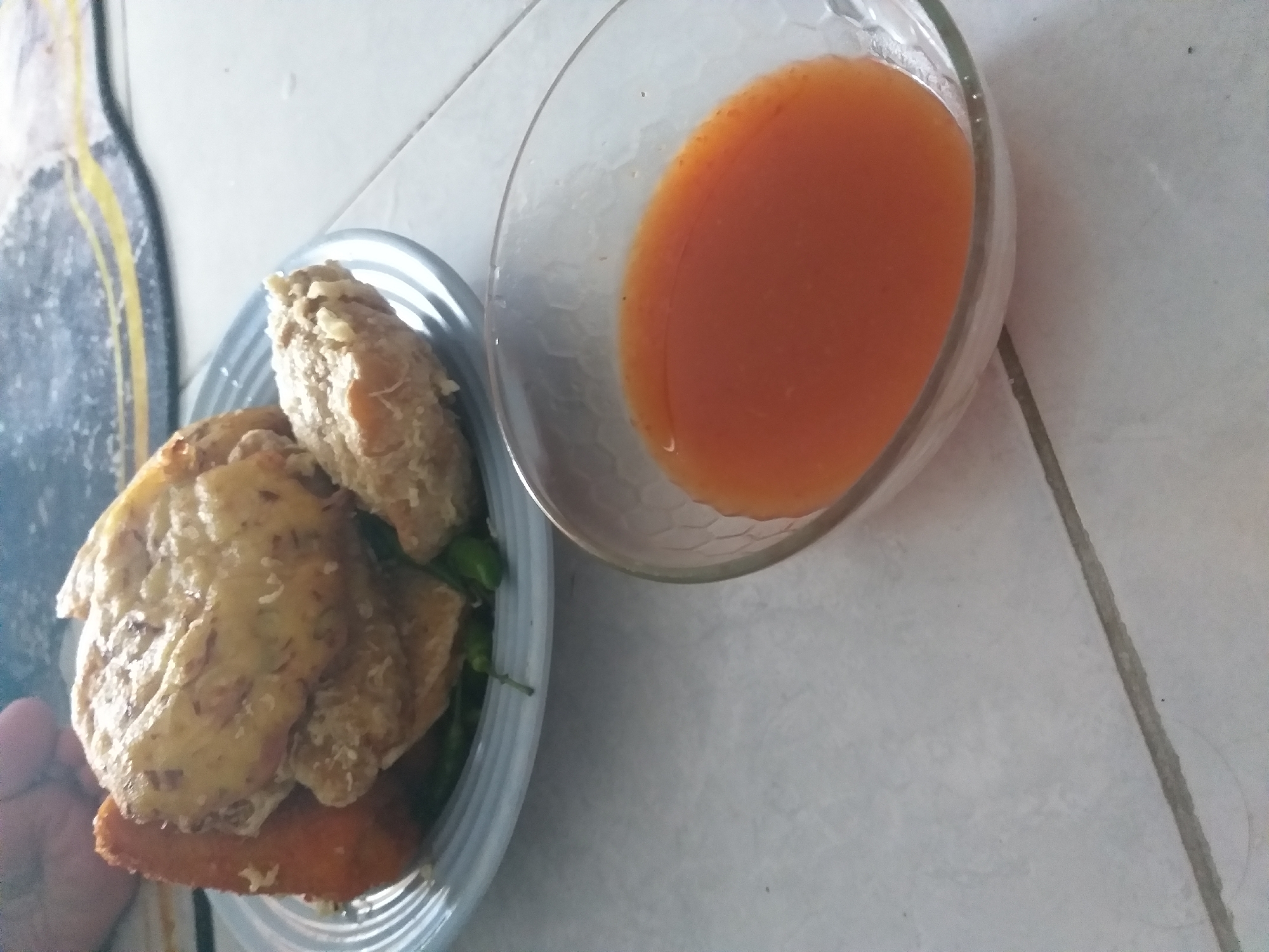 A close-up, slightly blurry, top-down shot shows a light blue plate with several fried food items, including what appears to be fried tofu and chicken, with some green chili peppers underneath. To the right of the plate is a clear glass bowl filled with a bright orange sauce. The plate and bowl rest on a tiled surface. The background is out of focus, but a textured mat with yellow and dark grey patterns is visible. The scene appears to be indoors, likely in a home setting in Air Jamban, Indonesia, given the context.
