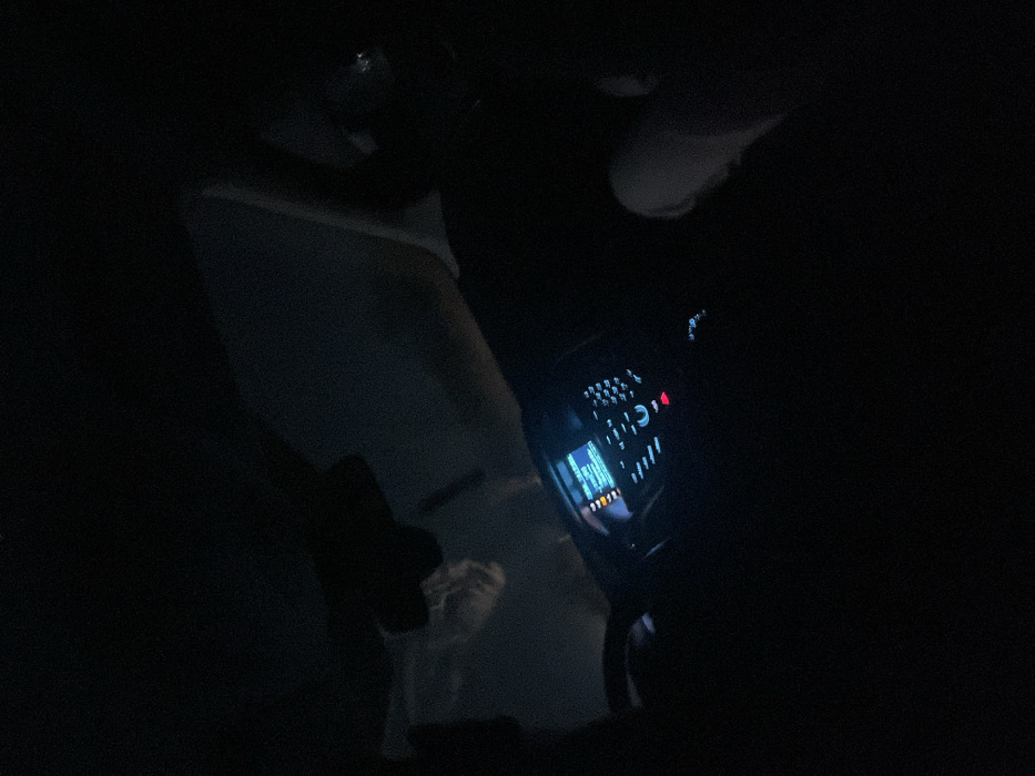 The image depicts the dark interior of a vehicle at night, located in Dalgo Pole, Bulgaria. The perspective is from the passenger side, looking towards the driver's area and dashboard. The dashboard's infotainment screen is illuminated in blue, displaying unreadable text, flanked by various controls with faint red and white indicator lights. A dark steering wheel is partially visible on the left. A rear-view mirror hangs from the ceiling, and beneath it, a faint, blurry, light-colored form resembling a human head or face is visible, possibly a reflection or an object. On the right side of the frame, the upper thigh and knee of a person, presumably a passenger, are visible, clad in light-colored clothing. The vehicle appears to be stationary or in motion with its electrical systems active.