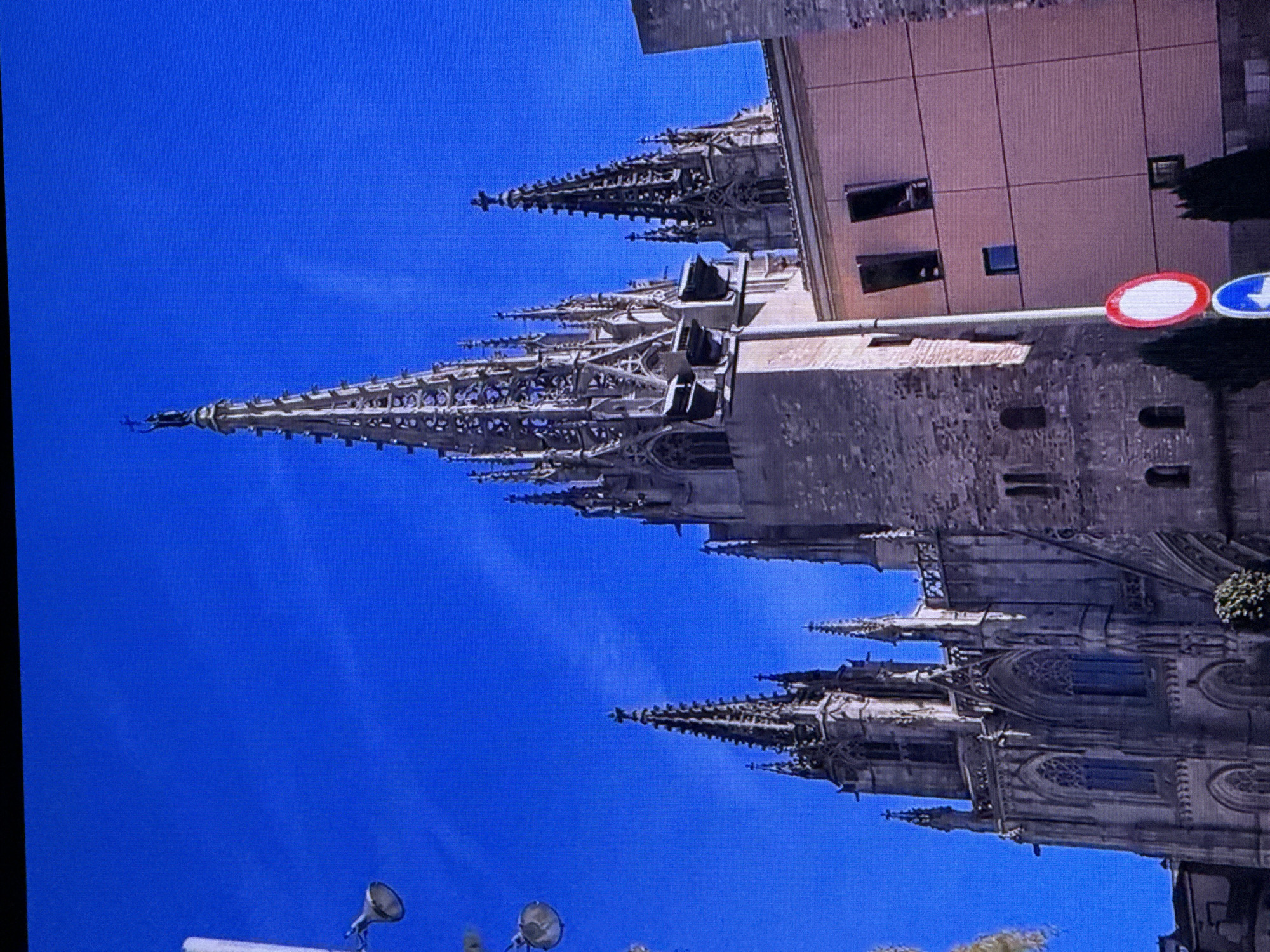 The image shows a Gothic-style cathedral with multiple spires reaching towards a bright blue sky. The architecture is ornate, with intricate stonework and pointed arches. Part of a modern building with a pink facade and several windows is visible to the right of the cathedral. There are also traffic signs, including a red and white circular sign and a blue directional sign, positioned on a pole in the foreground. In the lower part of the image, two light fixtures are visible, suggesting a street or public area. The scene appears to be set outdoors on a clear, sunny day. The orientation of the image is such that the cathedral and buildings are viewed from a low angle, emphasizing their height and grandeur. The presence of modern buildings and traffic signs alongside the historic cathedral hints at a mixed urban environment. There is no visible text in the image.