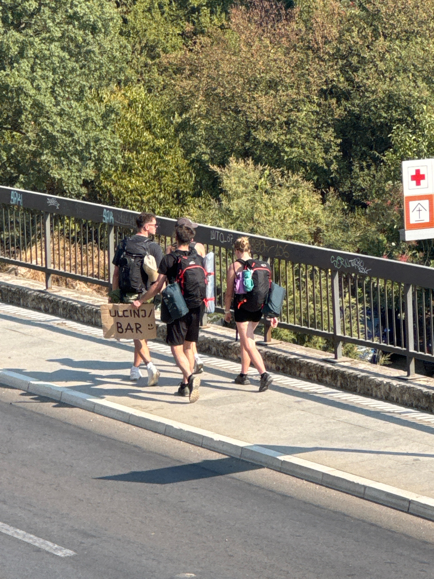 This is a full shot, taken from a high angle, of three individuals walking across a bridge in Boreti, Montenegro. Two individuals, appearing to be male, are walking side-by-side, with the one on the left carrying a cardboard sign that reads "ULCINJ/BAR :)". Both are wearing backpacks. The third individual, appearing to be female, is walking behind them, also wearing a backpack. The bridge has a metal railing with graffiti on it. The background is filled with trees and foliage. A red cross symbol and a pictogram of a house are visible on a signpost to the right of the bridge. The bridge overlooks a road with a curb and a white line marking.