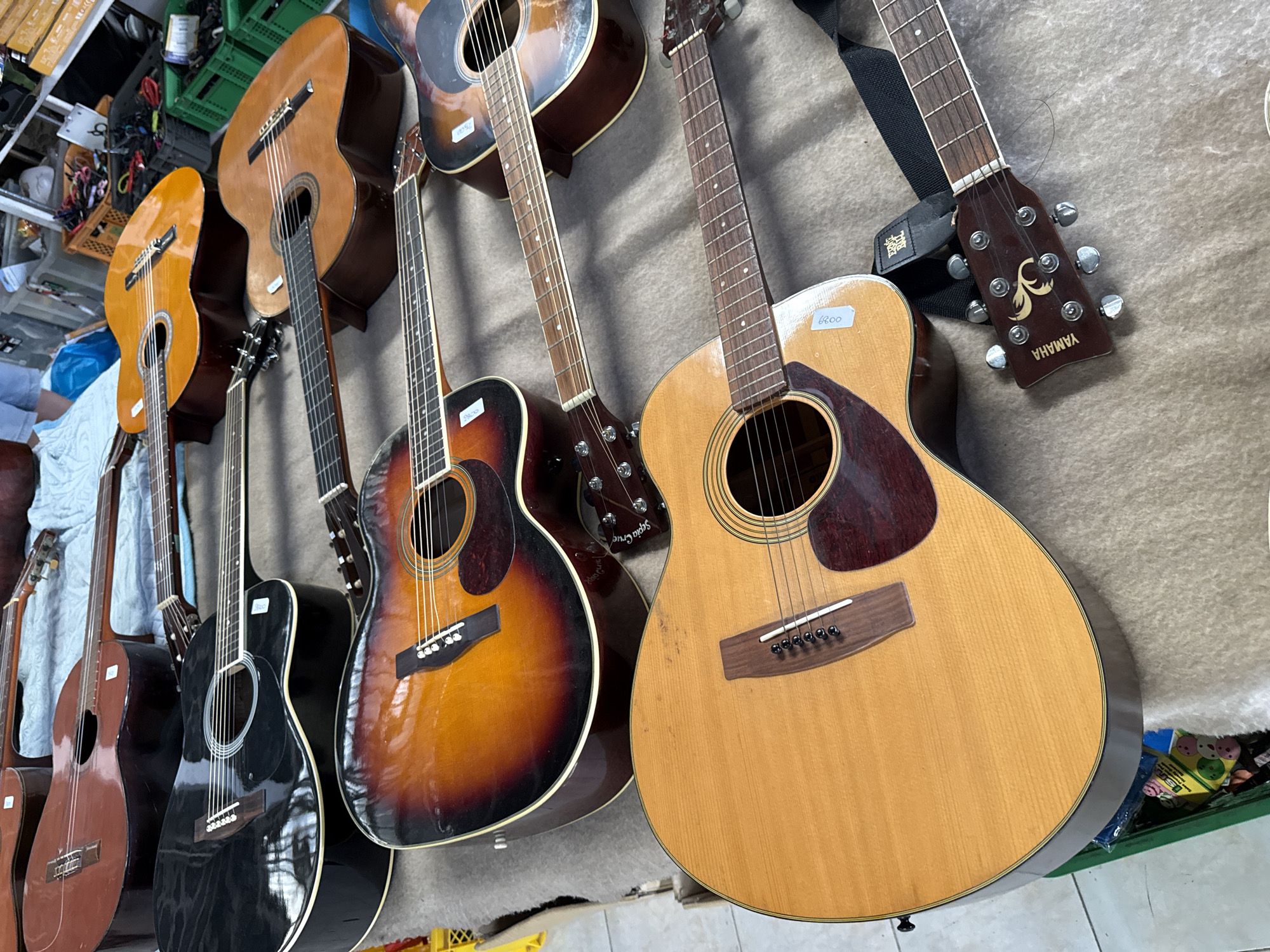This image shows a close-up, overhead view of several acoustic guitars laid out on a brown, textured surface, possibly a blanket or rug, in what appears to be an outdoor market. The guitars are positioned at various angles. In the foreground, a light-colored acoustic guitar with a natural wood finish is prominent. It has a dark red pickguard, silver tuning pegs, and a wooden bridge with white saddles. A sticker with the number "6000" is visible on the guitar's body. To the right of this guitar, the headstock of another guitar is visible, featuring the "YAMAHA" logo and a stylized "Y" emblem in gold, along with silver tuning pegs. A black guitar strap is attached to this headstock. Behind these guitars, other acoustic guitars are visible, including one with a sunburst finish (shades of orange, red, and brown) and a black acoustic guitar. Some of these guitars also have price tags or labels. The background is slightly out of focus but suggests a marketplace setting with glimpses of green plastic crates, a yellow shopping basket, and fabric. The overall impression is that of a display of guitars for sale. There are no people clearly visible in the frame. The location of Chalong, Thailand, is not directly evident from the image itself, but the description assumes this context.