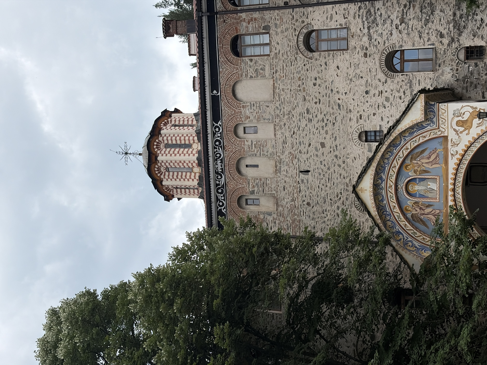The image shows a stone building with a red and white striped dome topped with a cross. The building has arched windows and ornate decorations. In the foreground, a large tree with green leaves partially obscures the view. The sky is overcast. The building is located in the Rila Monastery, Rilski manastir, Bulgaria.
