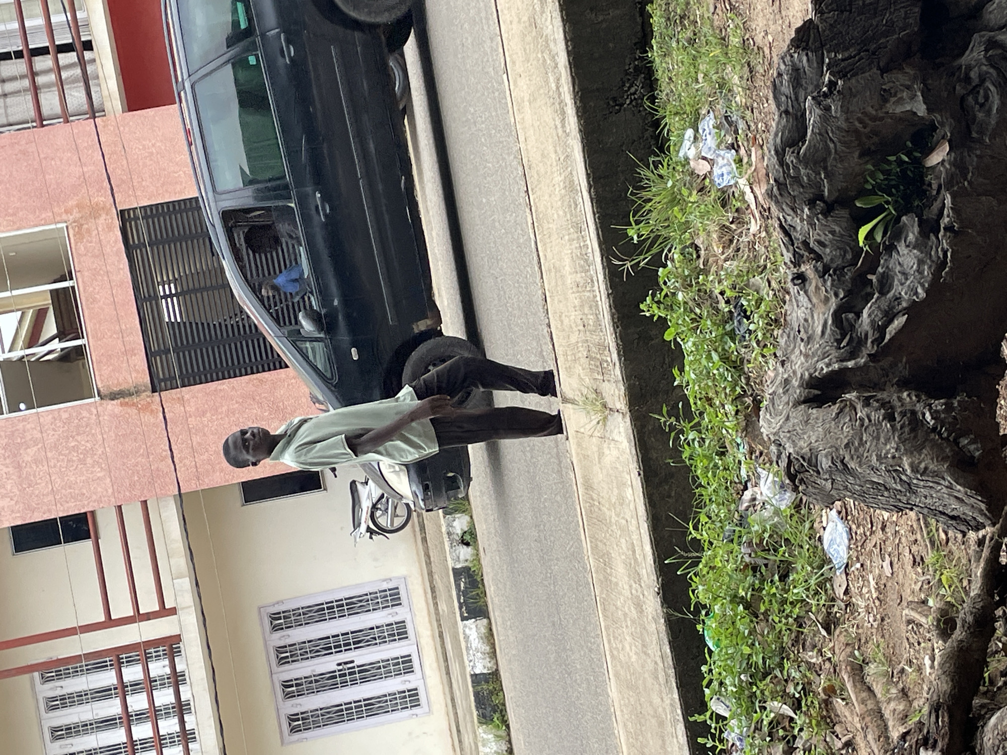 A young Black male wearing a light green collared shirt and dark pants stands on a concrete sidewalk beside a road. Behind him, a black minivan is driving away. The minivan has tinted windows and appears to be carrying at least one passenger. To the left of the minivan is a white motorcycle. In the background, there are buildings with multiple floors, featuring balconies and windows. The buildings are painted in light colors, with some having pink or red accents. The street is paved with asphalt, and there is a curb separating the road from the sidewalk. The foreground of the image shows a patch of overgrown green vegetation and tree roots, with some scattered litter. The scene appears to be in an urban environment, likely in Jos, Nigeria, given the architectural style and the general appearance of the surroundings.
