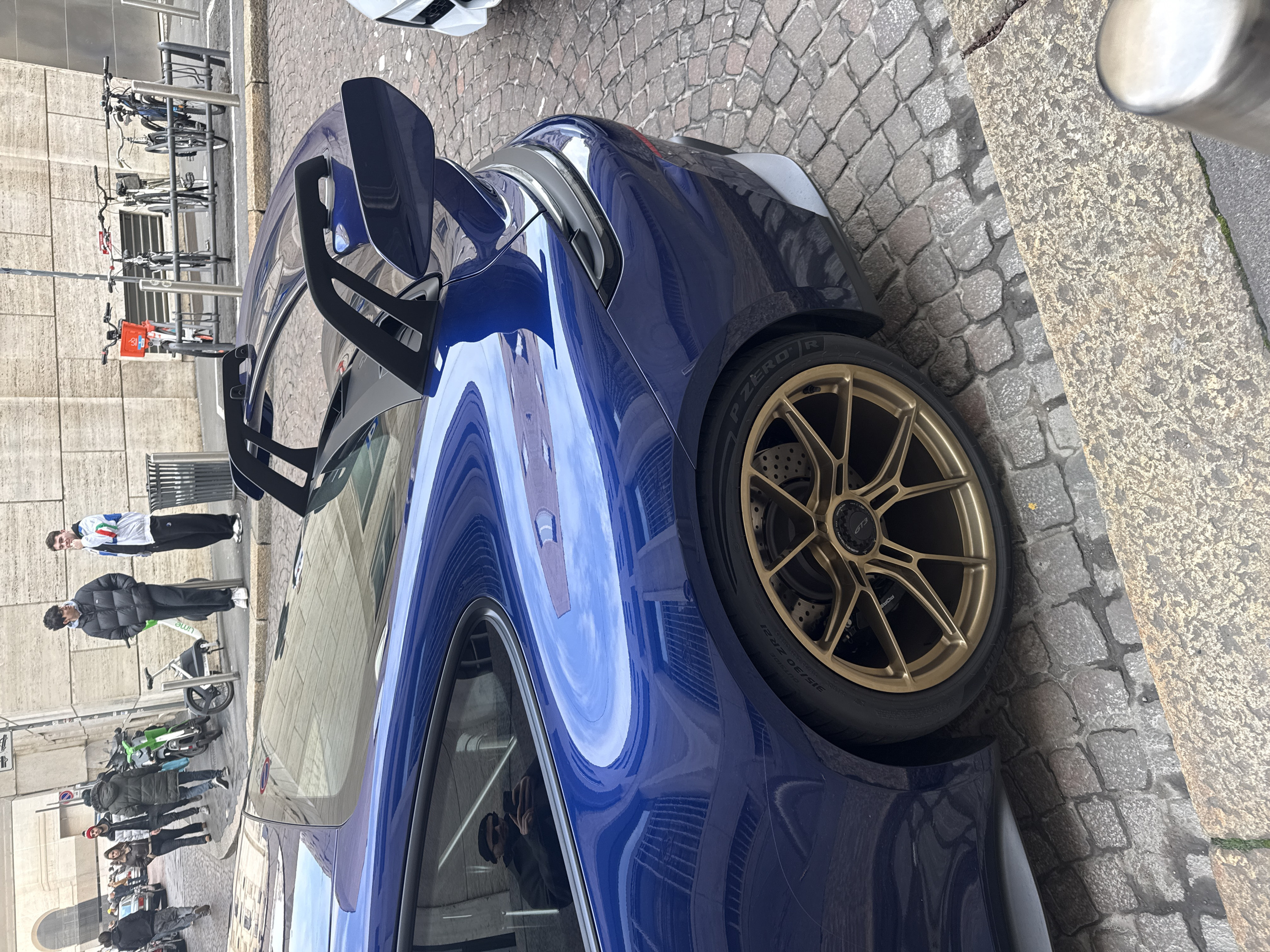 A dark blue Porsche 911 GT3 sports car is prominently parked on a cobblestone street in a historic, central urban area of Milan, Italy. The vehicle features large gold multi-spoke wheels with "GT3" center caps and Pirelli P Zero R tires (size 315/30 ZR21 on the rear). Perforated brake discs are visible behind the front wheel. A distinctive dark-colored rear wing with dual supports is present. A faint, thin scratch is visible on the car's rear fender above the wheel. The highly reflective paint of the car mirrors surrounding light-colored stone buildings, one of which displays prominent arched architectural features.
In the background, on the street and adjacent sidewalk, multiple pedestrians are present, dressed in winter clothing. On the far left, a person walks away from the camera. Further back, a couple approaches, with the man wearing a red beanie and the woman a brown puffer jacket. Near the center, a person in a dark green puffer jacket walks away. On the sidewalk to the right, two young men stand, one in a black puffer jacket and the other in a white jacket with red, white, and green stripes. A metallic trash can and several shared mobility devices, including green Lime scooters and bicycles (some parked in a metal rack, one with an orange basket), are situated on the sidewalk. A white vehicle is partially visible on the far right. Reflected in the rear passenger window of the Porsche, a man wearing a dark beanie and sunglasses holds a smartphone, appearing to take a photograph. The scene is illuminated by bright, overcast light.