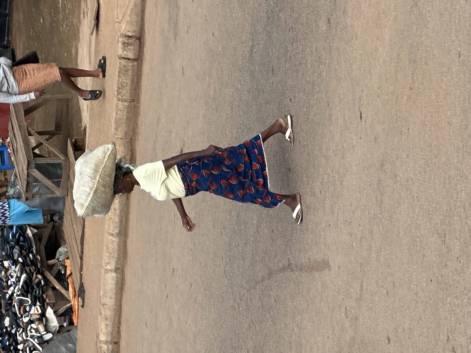 A woman walks across a paved street in Bukuru, Nigeria, carrying a large, irregularly shaped bundle on her head. She is wearing a light yellow t-shirt and a blue skirt with a red floral pattern, and white flip-flops. To the left of the woman, a stall is visible with a large pile of footwear, including many sandals and flip-flops. Behind this stall, a wooden structure is present, and further back, another individual is partially visible wearing a light-colored long-sleeved shirt and a peach-colored skirt. The street is made of asphalt, with a raised concrete curb along the side. The background shows a market-like setting with various wooden structures and stalls.