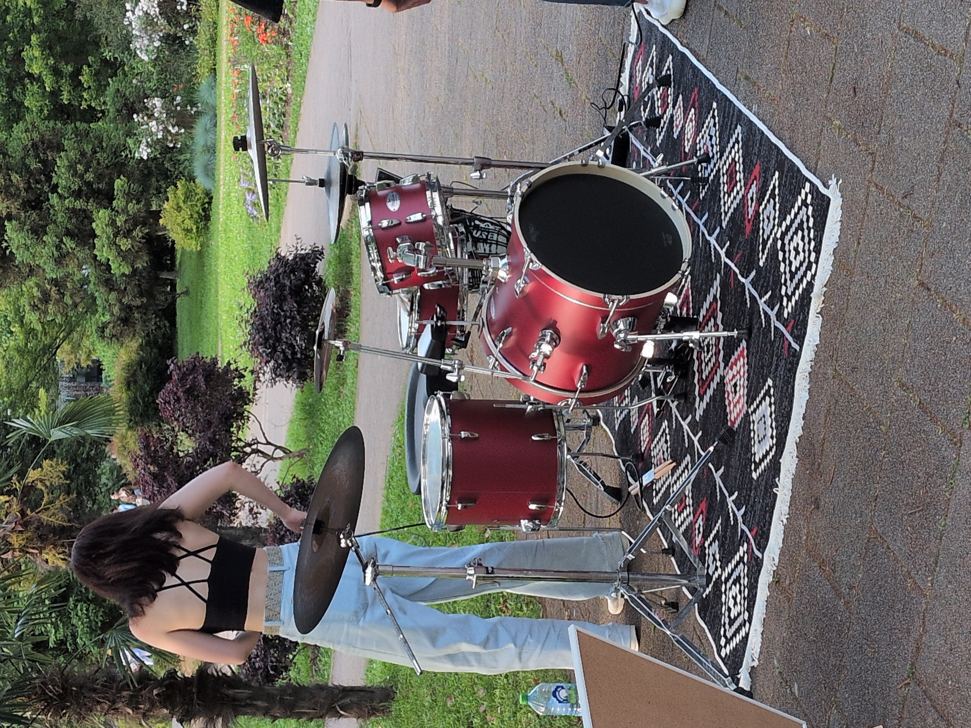 The image captures an outdoor scene in a park setting within the city of Sochi, Russia. A five-piece drum kit, featuring dark red shells with chrome hardware, is prominently displayed on a rectangular rug. The rug has a black background with white, red, and grey geometric diamond patterns and white fringed edges, placed on grey interlocking pavers. A person, viewed from the back, stands to the left of the drum kit. This individual has dark hair and is dressed in a black cross-strap crop top, a patterned light-colored belt, and light blue wide-leg jeans. Their arms are bent with hands near the waist. A crash cymbal is mounted on a stand directly in front of this person, with a hi-hat cymbal positioned to its right. A clear plastic water bottle with a blue and white label is on the ground to the far left, adjacent to a partial view of a light-colored triangular board. Two drumsticks are visible lying on the rug near the bass drum. The lower leg of another person, wearing dark trousers and a white sneaker, is partially visible on the far right. The background is dominated by lush green vegetation, including tall palm trees on the left, various green and dark red-leafed bushes, and a paved pathway leading into the distance where more trees and white flowering shrubs are visible. The environment suggests a public garden or park.