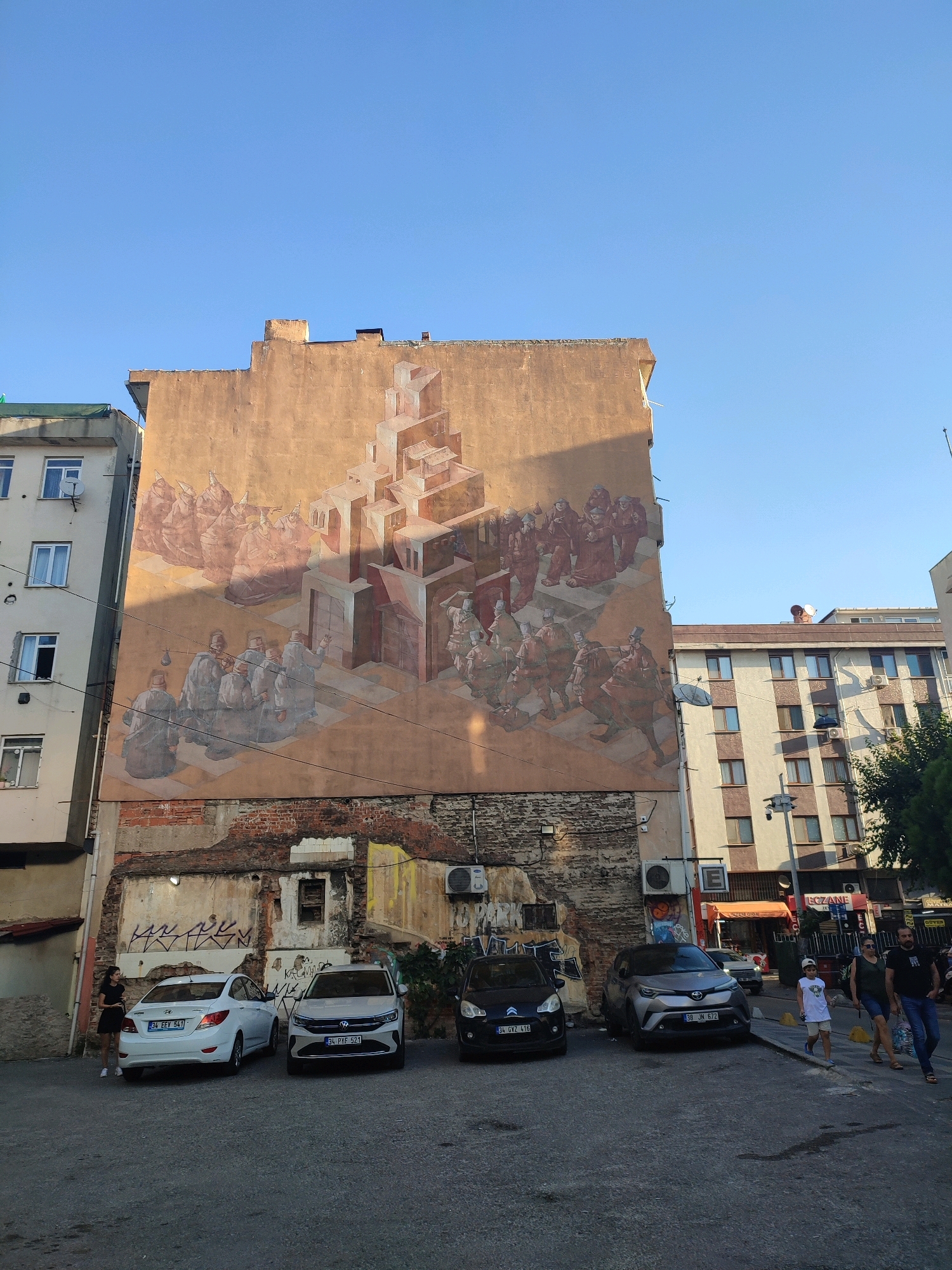 A wide-angle, low-angle shot depicts a mural on the side of a tan-colored building in Kadıköy, Turkey. The mural features a surreal, Escher-esque architectural design with figures in historical-style clothing. The building itself is weathered, with exposed brick and graffiti on the lower sections. In front of the mural, several cars are parked in a lot. To the left of the mural is a modern, white apartment building. To the right, a multi-story building with a checkered facade dominates the background. People are visible walking on the sidewalk and near the parked cars. The sky is clear and blue.