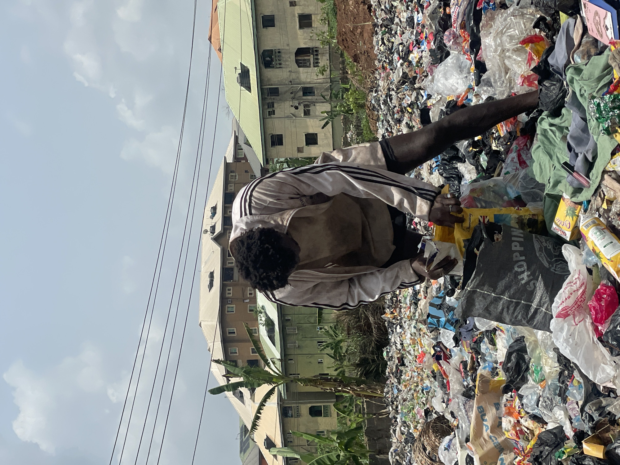 An individual is positioned in a large pile of garbage in Onitsha, Nigeria. The individual wears a white tracksuit with black stripes and short black hair. The person is leaning over the garbage, handling a yellow plastic container. The garbage comprises various materials, including plastic bags, clothing, and food containers. In the background, buildings and utility lines are visible. The sky is partially cloudy. Palm trees are growing near the garbage pile.