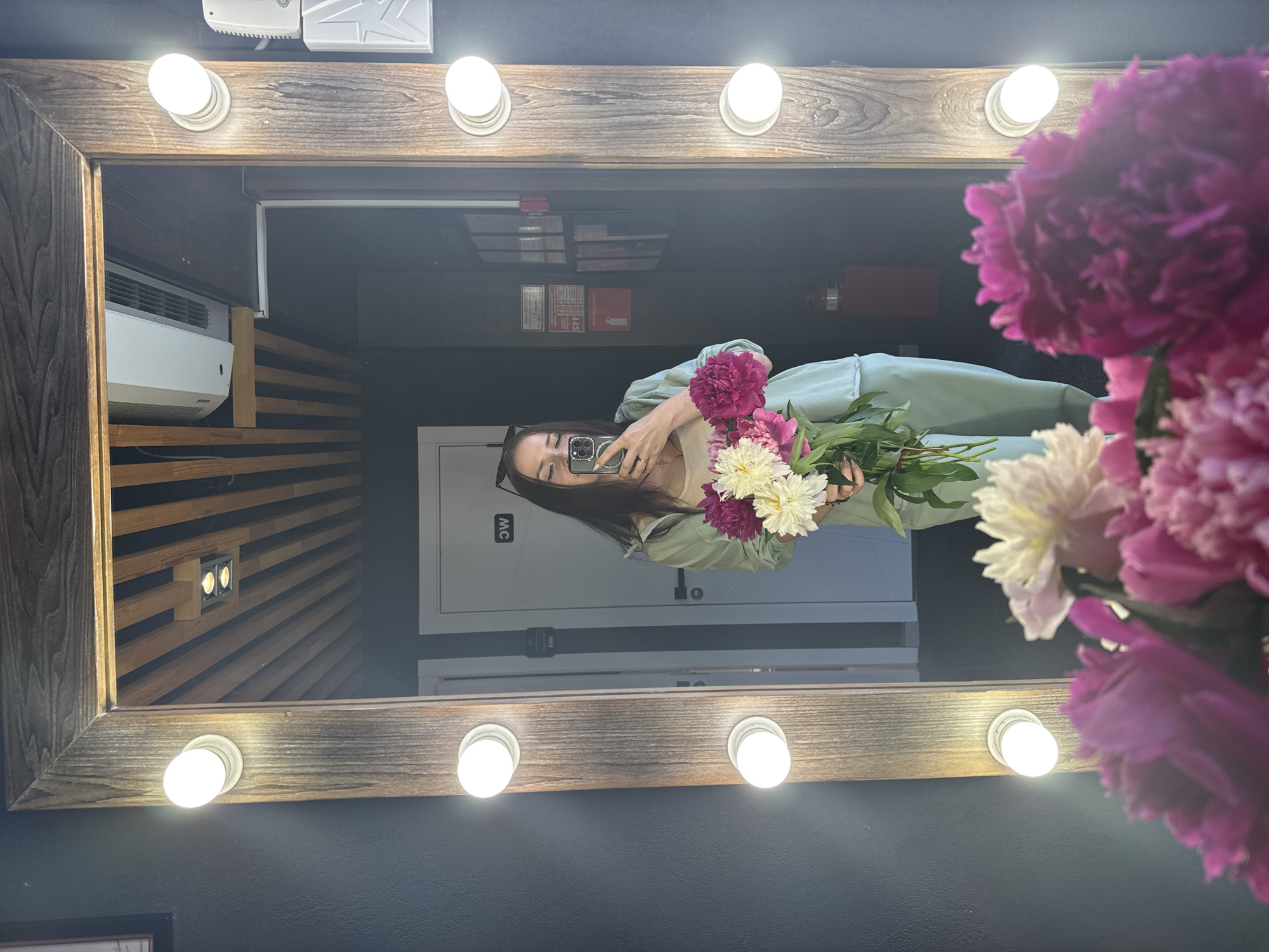 A young woman with long dark hair, wearing a light green loungewear set and a light pink top, stands in front of a large, rustic wooden framed mirror. She is holding a bouquet of peonies in various shades of pink and white, and taking a selfie with her phone. The mirror is illuminated by several round light bulbs along its sides. In the reflection, the woman is positioned in what appears to be a room with a dark wall on the left and a white door on the right, marked with a "WC" symbol. The ceiling is visible in the reflection, featuring wooden slats and recessed lighting. Additional objects visible in the reflection include what appears to be an air conditioning unit mounted on the ceiling and some posters or signs on the wall. In the foreground of the image, out of focus, are more peonies. The scene is likely indoors, possibly in a dressing room or a makeup studio. There is no specific indication of the city of Chaikovsky, Russia within the image itself.