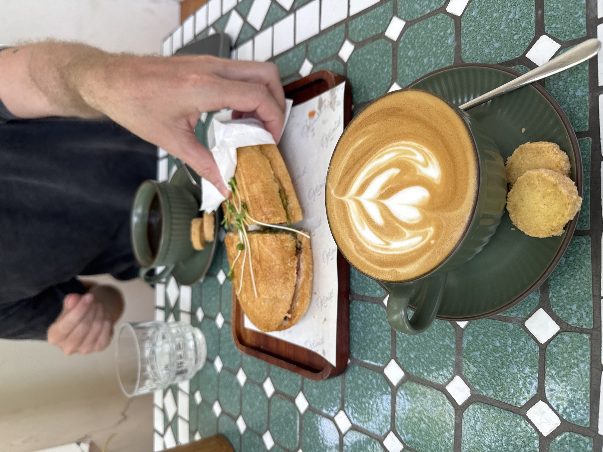 A person reaches for a sandwich on a wooden tray next to a cup of coffee with latte art and two cookies on a saucer. There is a small cup of coffee and a glass of water on the green, white, and black tiled table. The location is Unknown, Unknown.