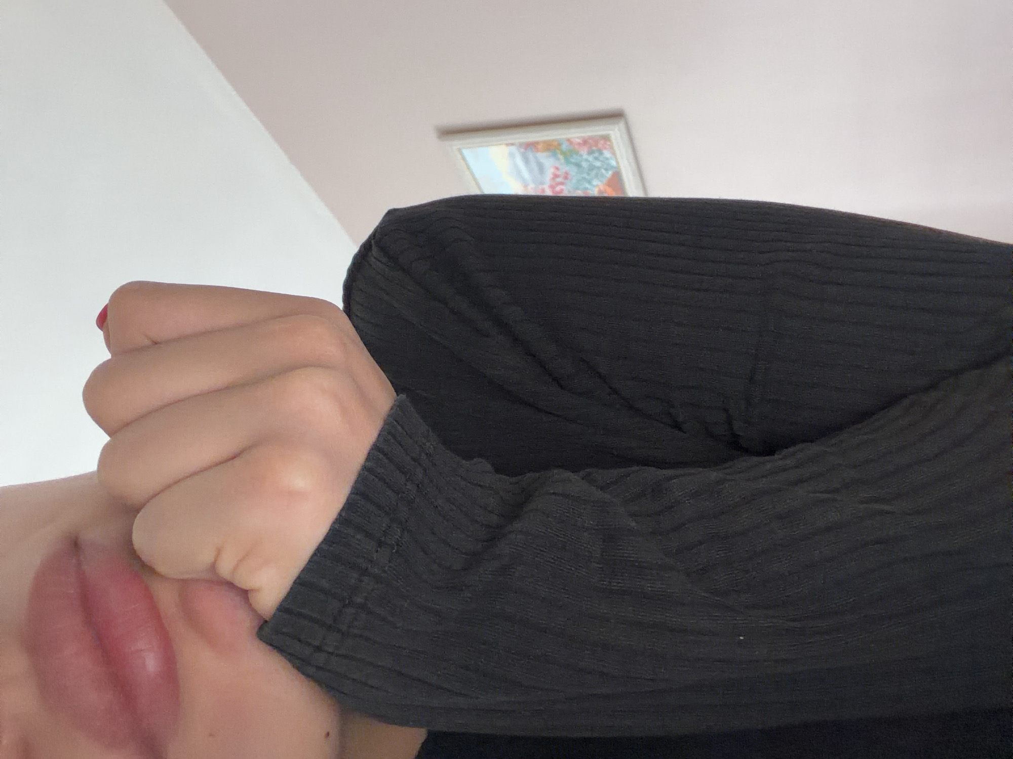 The image depicts a close-up, angled view of a person's face and upper torso. The person is wearing a black, ribbed long-sleeved garment. Their lips are visible, with a portion of their cheek and hand obscuring the rest of their face. The hand is partially clenched into a fist. In the background, a white wall and ceiling are visible, with a framed picture hanging on the wall. The location is not discernible from the image.