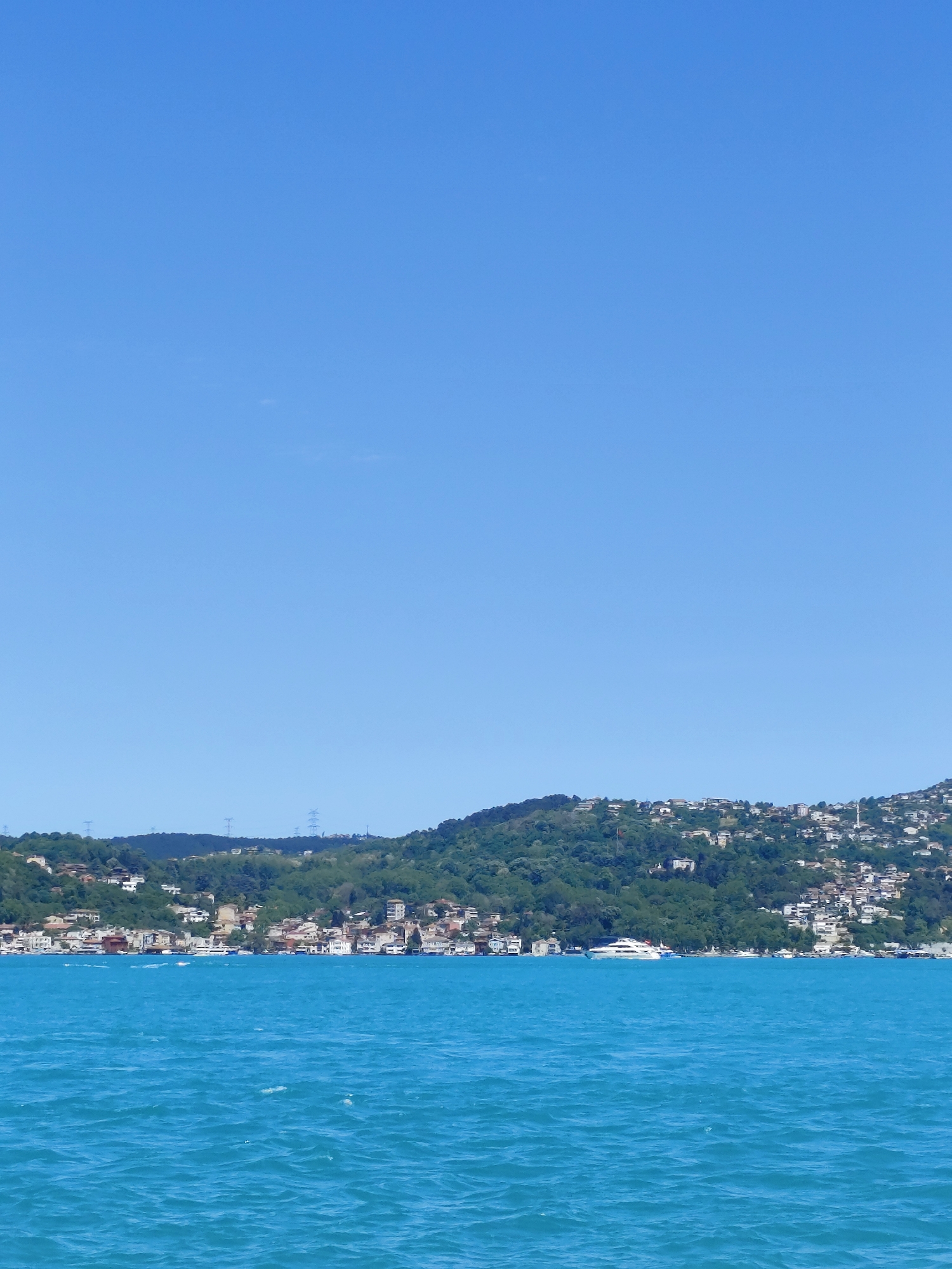 The image depicts a body of water, possibly the Bosporus Strait, with a shoreline visible in the mid-ground. The water is turquoise, indicating a clear day.
Along the shoreline, a hillside rises, covered in green vegetation interspersed with buildings, suggesting a residential or urban area built into the landscape. The style of buildings appears to be residential, and the layout suggests the photograph was taken in the Sarıyer district of Istanbul, Turkey.
A prominent white yacht sits on the body of water, visible.
The sky is a clear, cloudless blue, taking up the majority of the frame.