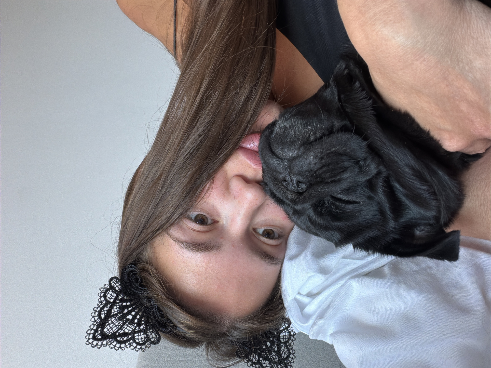 A young woman with long brown hair and a black lace headband resembling cat ears rests her head on a white pillow. She is embracing a black pug. The woman is looking at the camera with wide brown eyes. The pug's face is nestled against the woman's cheek. The scene appears to be indoors, possibly in a bedroom. The background is a light-colored wall.