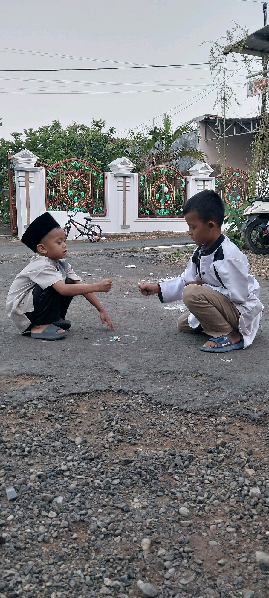 Two young boys are playing marbles on a cracked asphalt road in Keling, Indonesia. The boy on the left is wearing a black peci cap and a light-colored tunic, kneeling with his fist clenched. The boy on the right is wearing a white tunic with black accents and khaki pants, also kneeling with his fist clenched. In front of them, a circle is drawn on the ground with marbles inside. In the background, a white and brown fence with decorative metalwork, green foliage, a bicycle, and a motorcycle are visible. The sky is overcast.