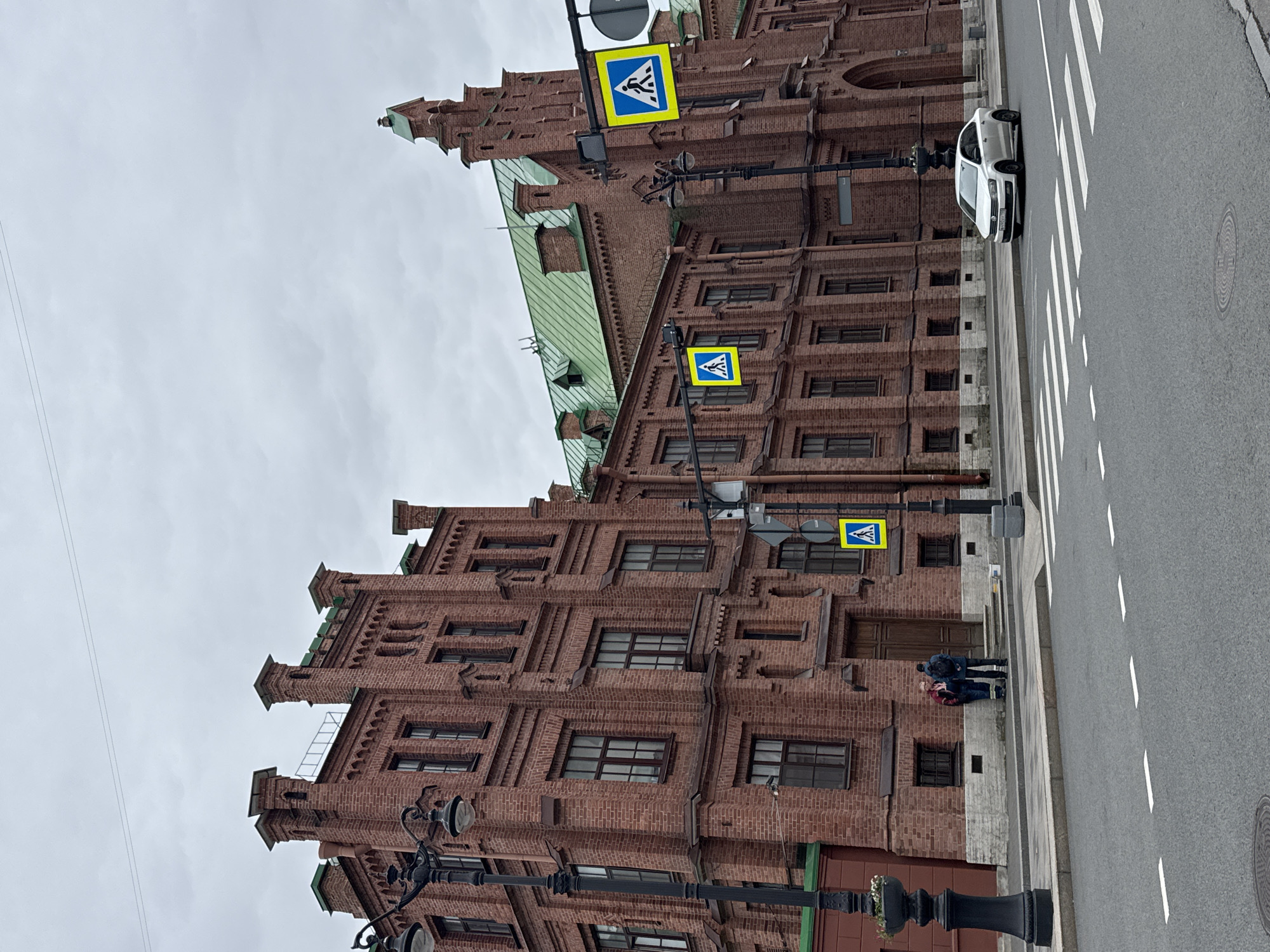 Here is a detailed description of the image:
**Setting:** An urban street scene in Saint Petersburg, Russia. The weather appears overcast.
**Building:** A large, ornate brick building dominates the view. The architecture is distinctive, with multiple tiers, many windows, and decorative brickwork. It has a green roof section.
**Street Scene:**
* A pedestrian crosswalk (zebra crossing) is visible on the right side of the frame.
* A white car is parked along the edge of the crosswalk.
* Three pedestrian crossing signs are visible, attached to lamp posts near the building.
* There are two people, dressed in winter clothes, standing on the sidewalk in front of the building.
* Elaborate black lampposts line the street.
* A set of power lines are visible against the grey sky.