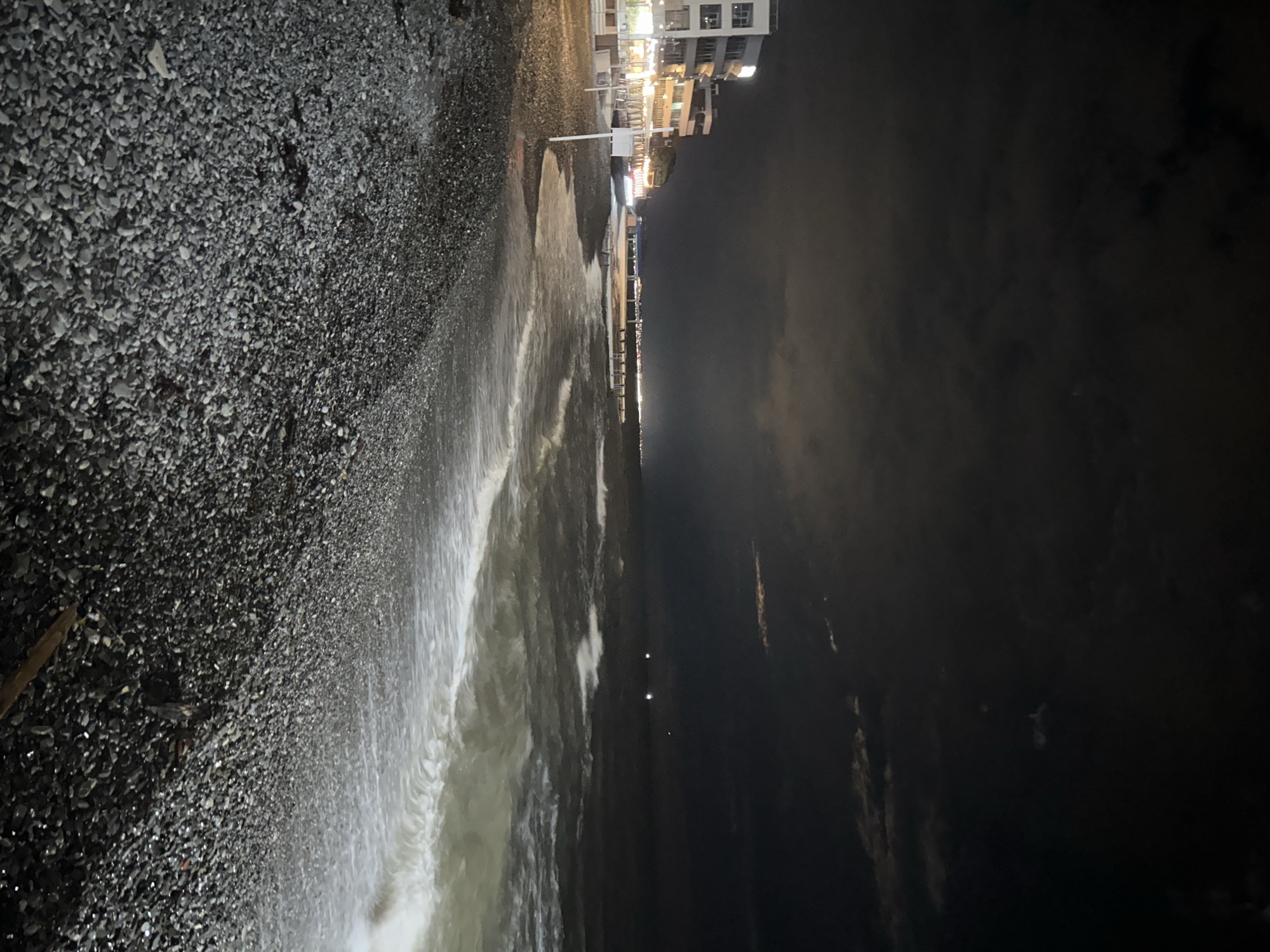 The image shows a nighttime view of a pebble beach at Sochi, Russia. The beach slopes from left to right, and is covered in dark grey pebbles. The sea crashes onto the beach with foamy white waves. Beyond the water line, the view fades into dark sky and clouds. In the distance, along the coastline, there are buildings illuminated by streetlights, suggesting a populated area. There are no people visible in the image. A wooden object is visible on the pebble beach on the left side of the frame.