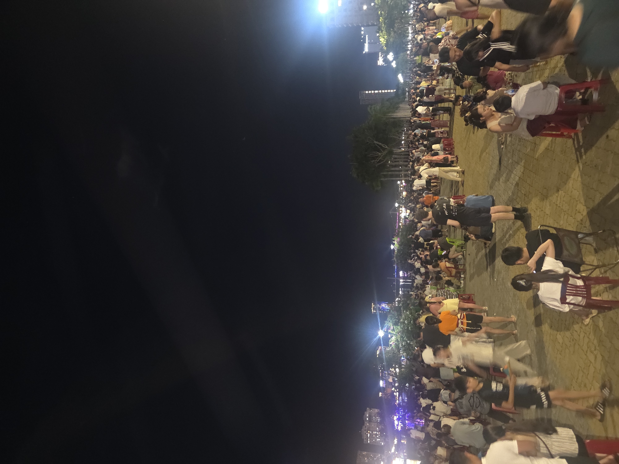 The image shows a crowded scene at night in Đà Nẵng, Vietnam. People are gathered along what appears to be a waterfront promenade. Many are seated on small plastic stools, while others are standing. The atmosphere is festive, with bright lights illuminating the area and buildings in the background. The crowd is densely packed, and individuals are interacting with each other. The background shows the city skyline with tall buildings. The surface the crowd is on appears to be paved.