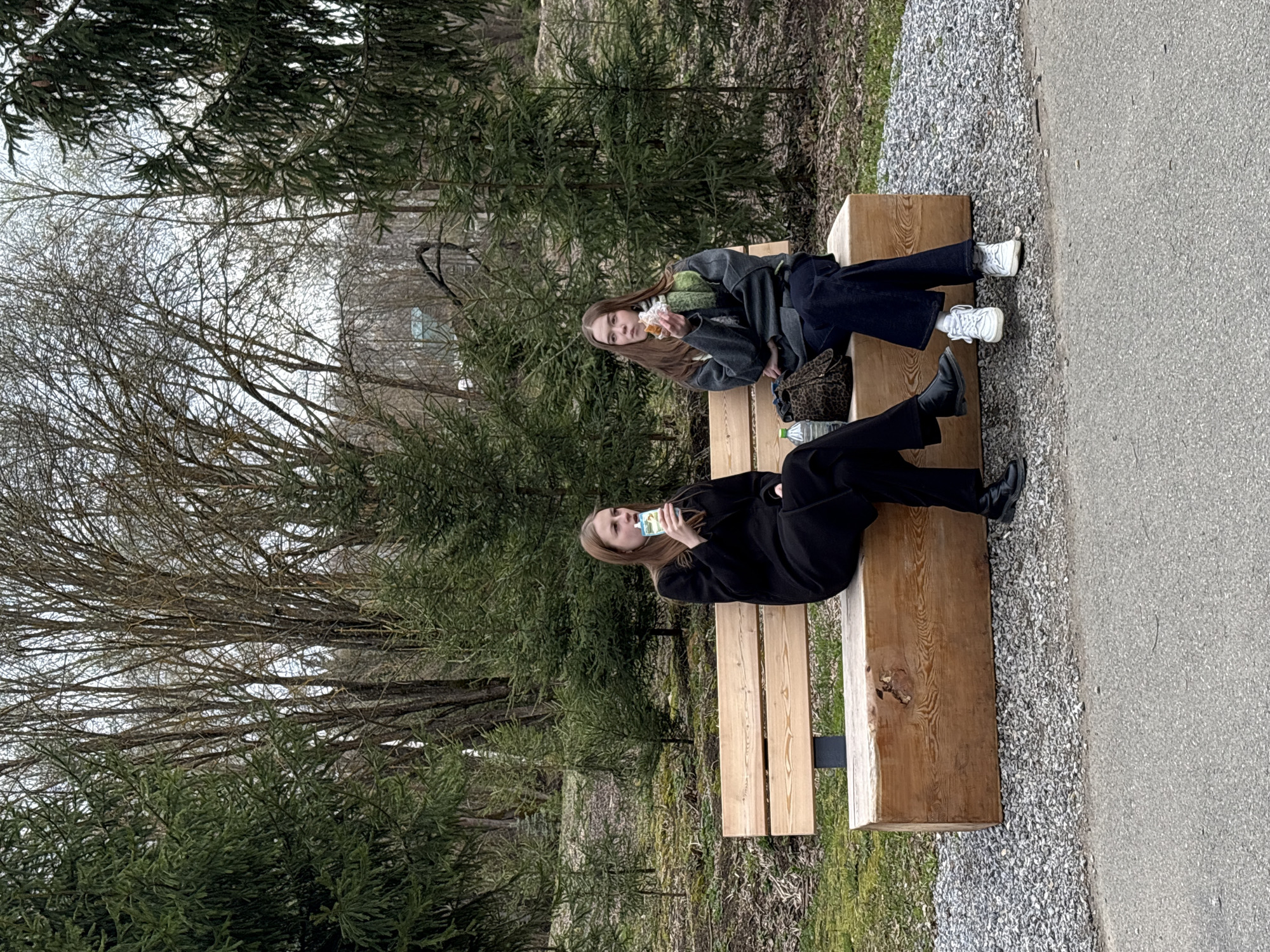 Two young women are seated on a wooden bench in a park-like setting with mature trees and a gravel path in Sdt Shcherbakovka, Russia. One woman is wearing a dark coat and trousers with black shoes and holding a juice box. The other is wearing a dark jacket with a green scarf and denim trousers with white sneakers, and is holding a sandwich. A purse and a plastic water bottle are placed on the bench.