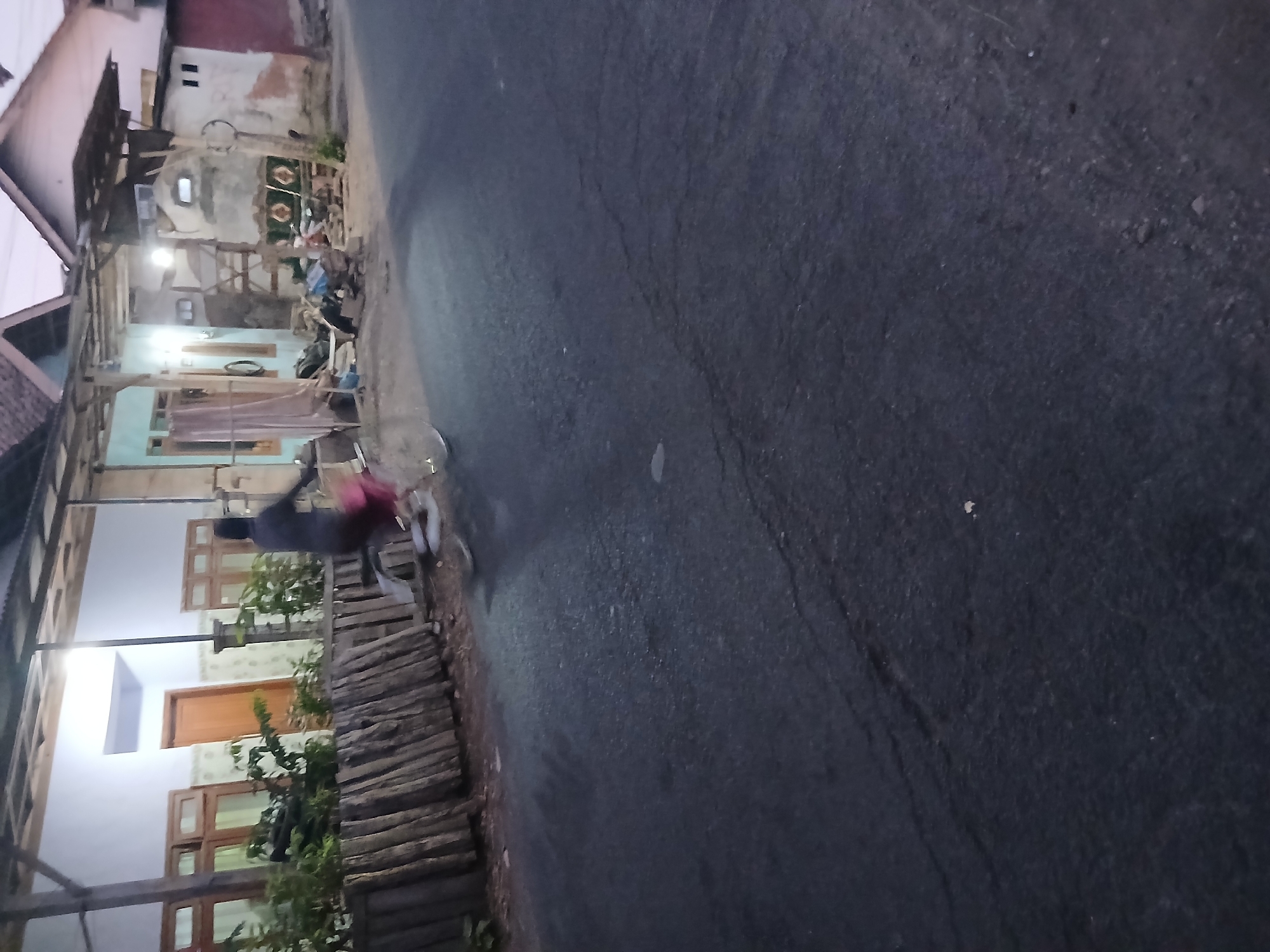 In Kademangan Kulon, Indonesia, at dusk, a person is cycling on an asphalt road. The rider, wearing dark clothing and a red skirt, is blurred due to motion. They are cycling past a row of wooden posts and then past a building with a white facade, wooden window frames, and a light blue section. The building appears to be a residential house with an overhang roof. Further down the street, another structure with an irregular facade and a darker roof is visible. The ground in front of the buildings is a mixture of dirt and gravel, with scattered debris. The lighting suggests it is either early morning or late evening, with artificial light emanating from the buildings.